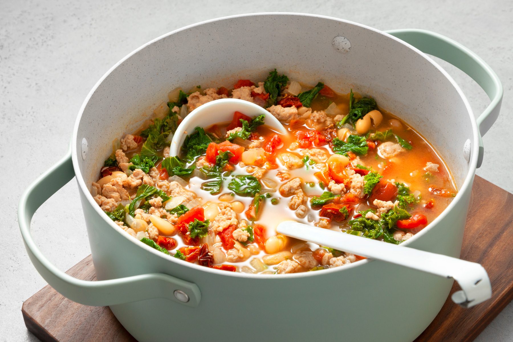 Table view shot of Italian Sausage Kale Soup; in a large skillet; serving spoon; wooden board; grey marble surface
