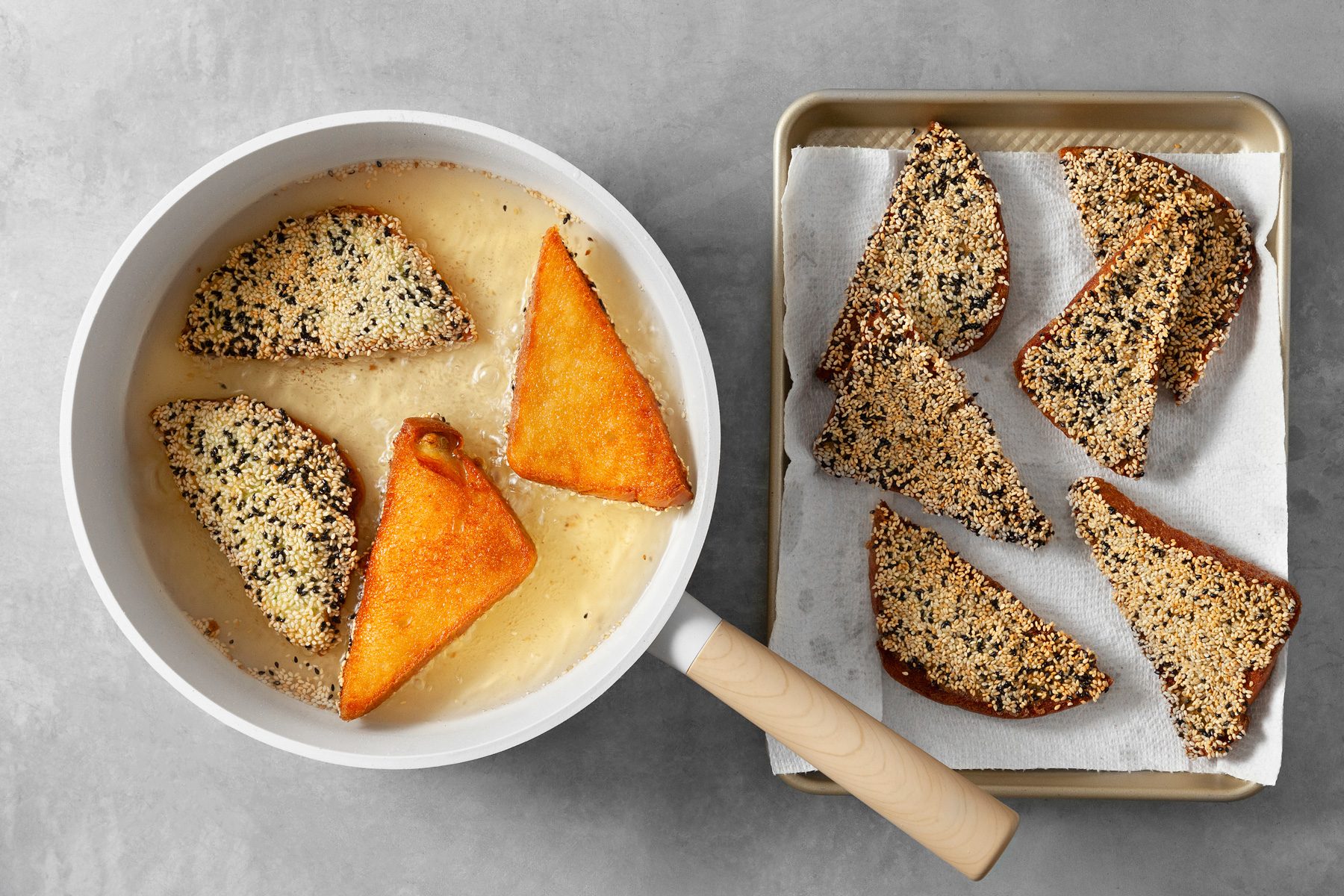 overhead shot; light grey background; Triangles of bread coated in a shrimp mixture and sesame seeds are being fried in a white pan filled with hot oil; The bread is turning golden brown and crispy; A paper towel-lined baking sheet is ready to receive the fried shrimp toast