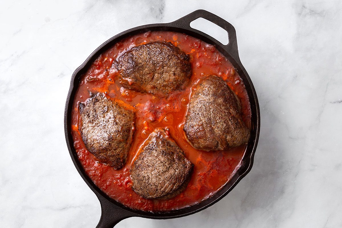 Top Sirloin steaks simmering with sauce in a large skillet for step five of Steak Pizzaiola recipe for Taste of Home