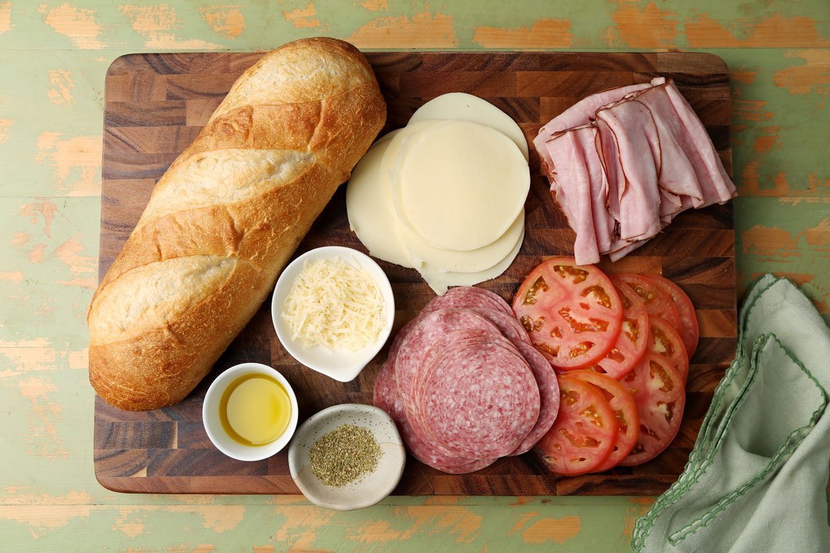 Ingredients for Taste of Home's Italian Sub laid out on a wooden cutting board on a green surface.