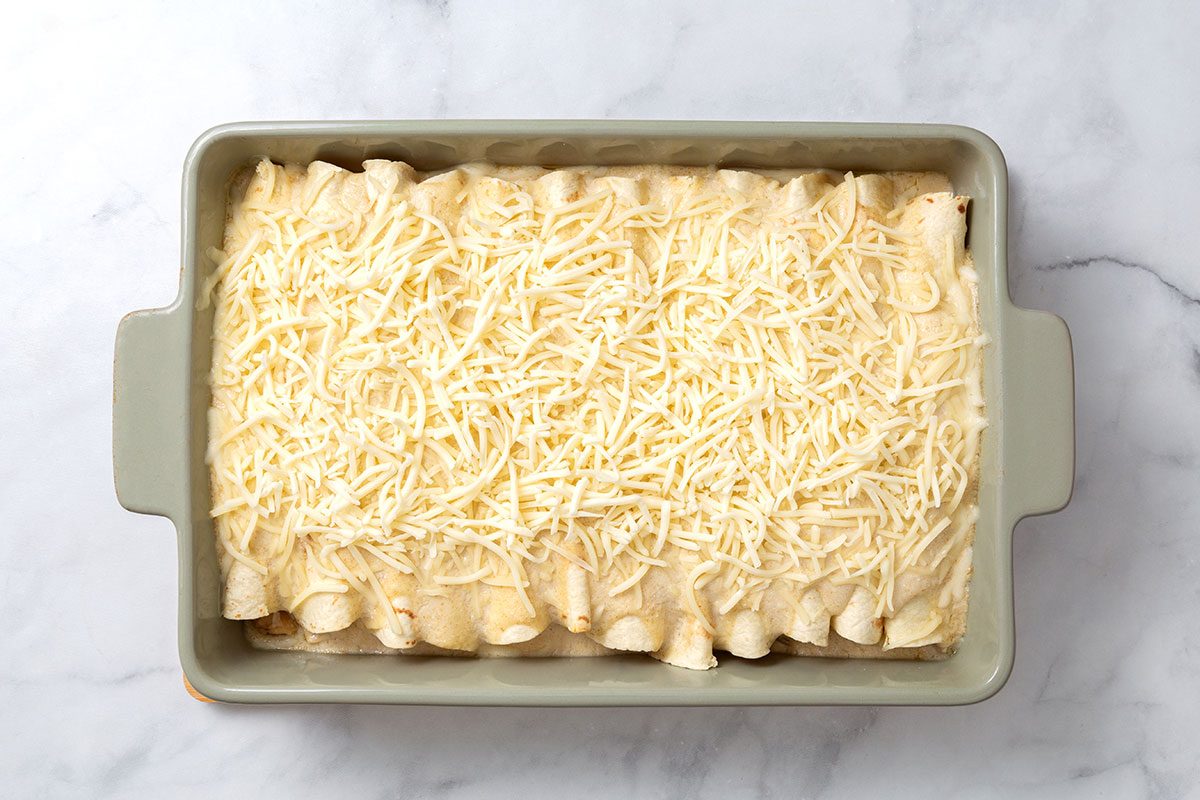 Rolled up tortillas in baking dish layered with white sauce and Monterey Jack shredded cheese