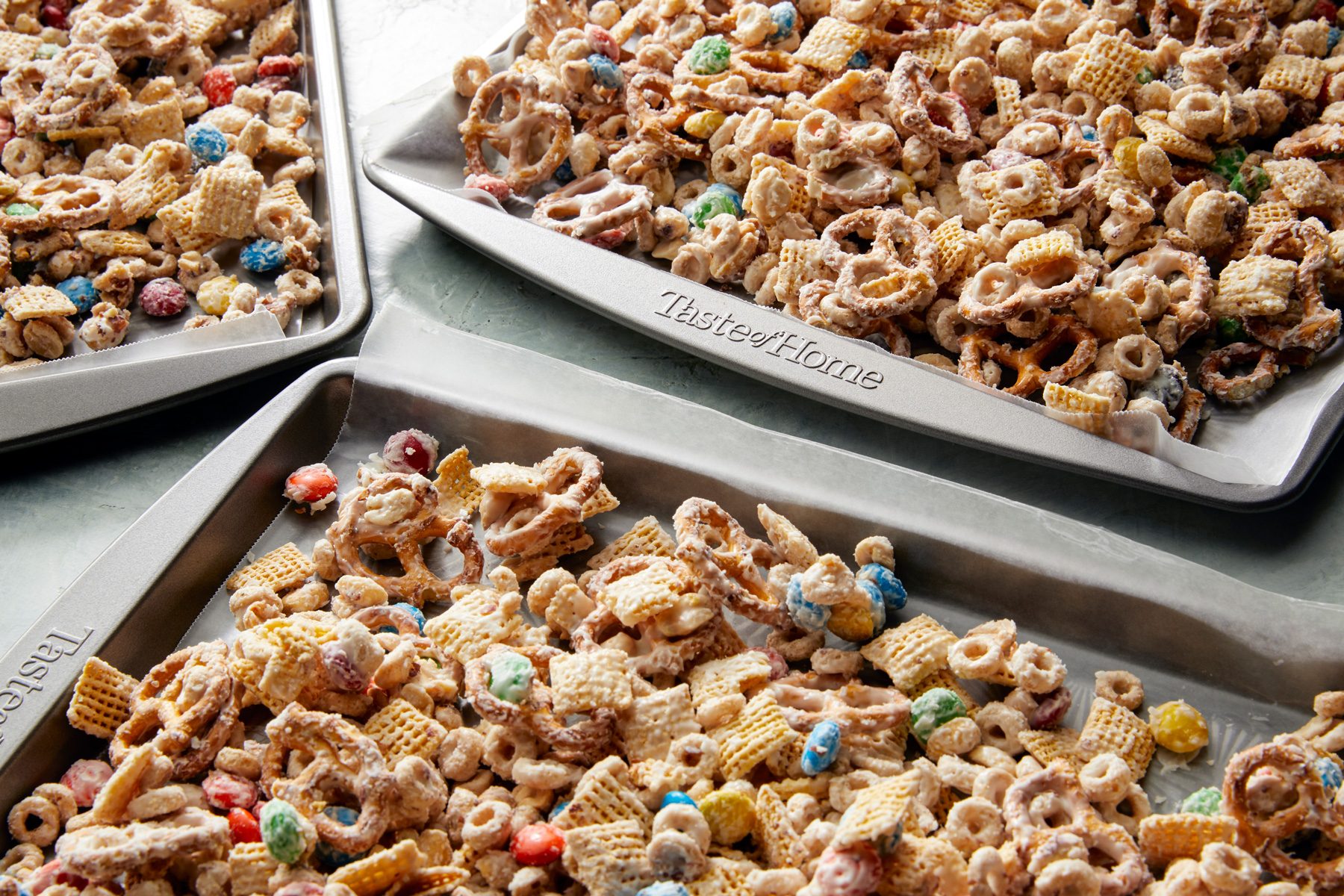 3/4th shot; white background; Three baking sheets are filled with a mixture of pretzels, Chex cereal, and colorful candy, all coated in a white chocolate mixture; The mixture is spread out evenly on the sheets, ready to be baked