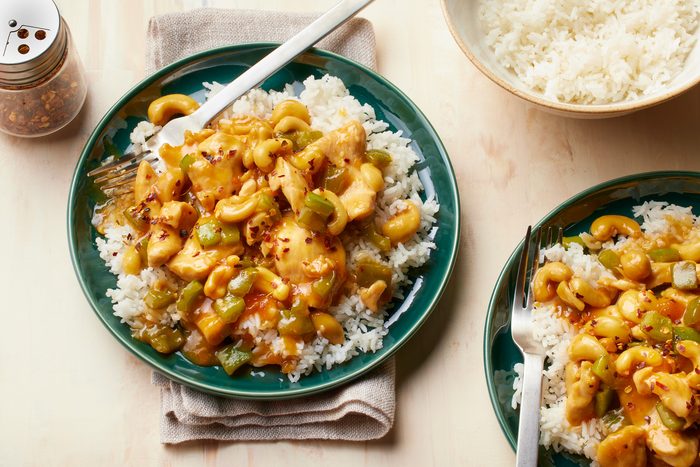 overhead shot of two plates of a dish called Apricot Chicken placed over wooden background, The dish consists of stir-fried chicken coated in a sweet and tangy apricot sauce, The chicken is served over a bed of white rice and garnished with red pepper flakes, The dish is accompanied by a bowl of steamed rice and a shaker of red pepper flakes
