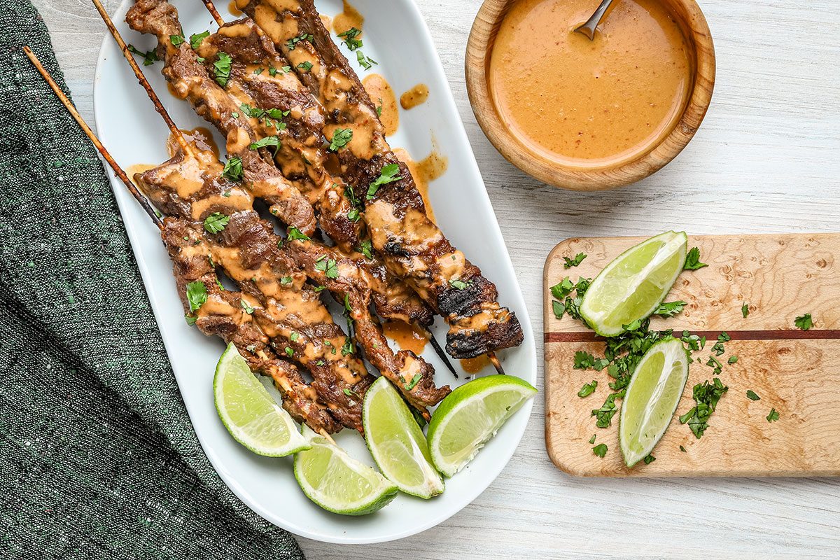 Beef satay skewers with peanut sauce drizzled over them along with lime wedges and cilantro.