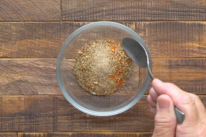 A person holding a spoon is mixing a variety of dried spices in a clear glass bowl. The bowl is placed on a wooden surface.