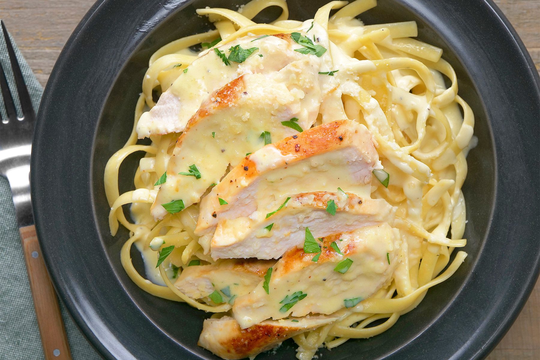 overhead shot of a plate of Chicken Alfredo is showcased, featuring tender pieces of grilled chicken nestled in a creamy, buttery Alfredo sauce, Fettuccine pasta forms the base of the dish, a silver fork is placed near the plate over kitchen napkin;