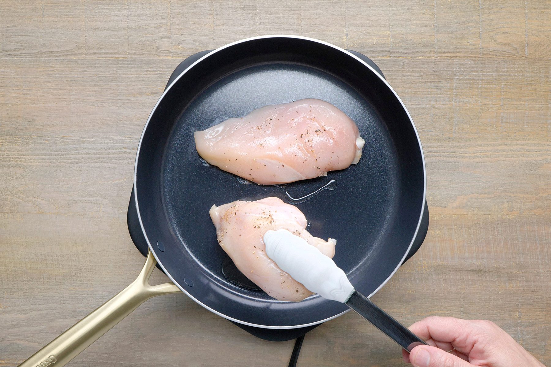 overhead shot of two chicken breasts cooking in a black skillet, The chicken breasts are a pale white color and are seasoned with black pepper, A spatula is being used to flip one of the chicken breasts;