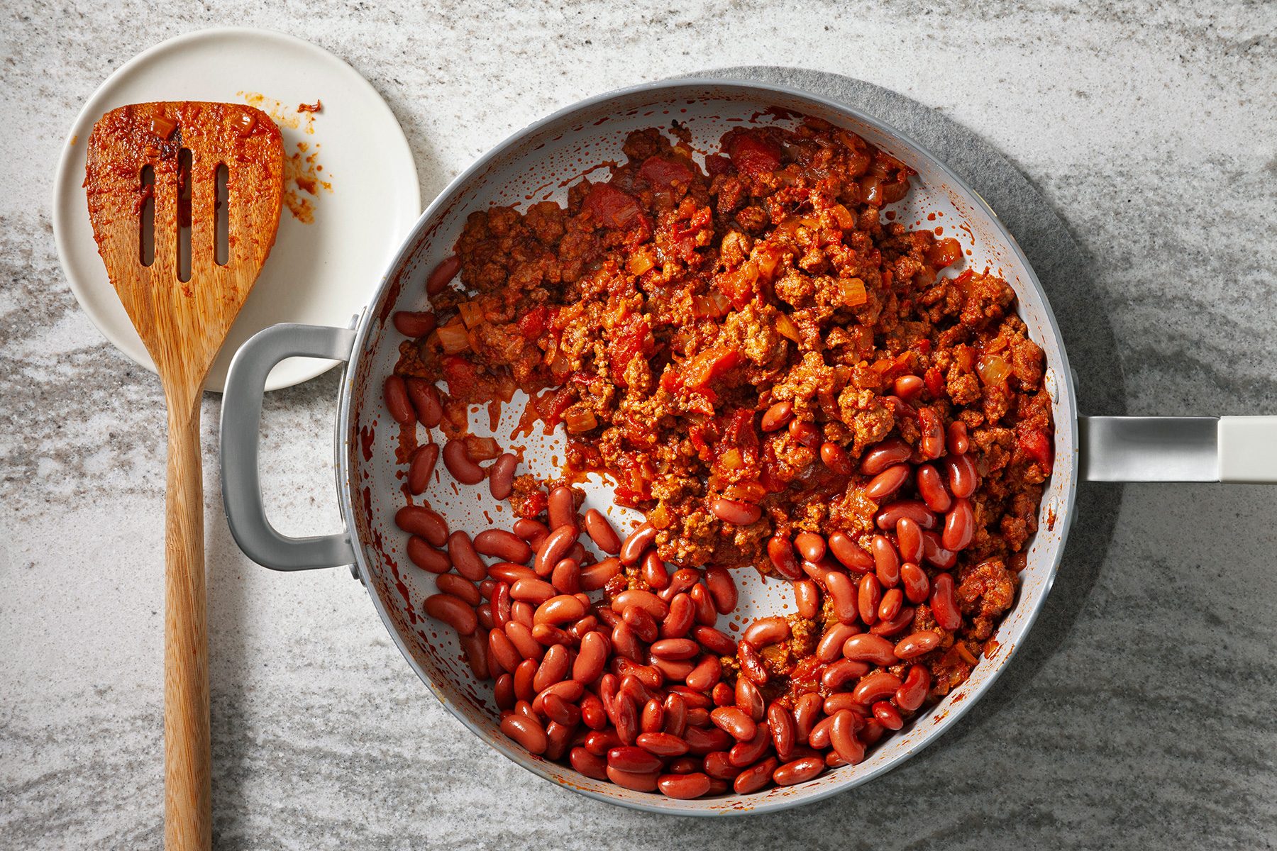 A skillet containing cooked ground meat and kidney beans in a red sauce sits on a gray counter. A wooden spatula with sauce rests on a small white plate next to the skillet, with remnants of sauce visible on it.