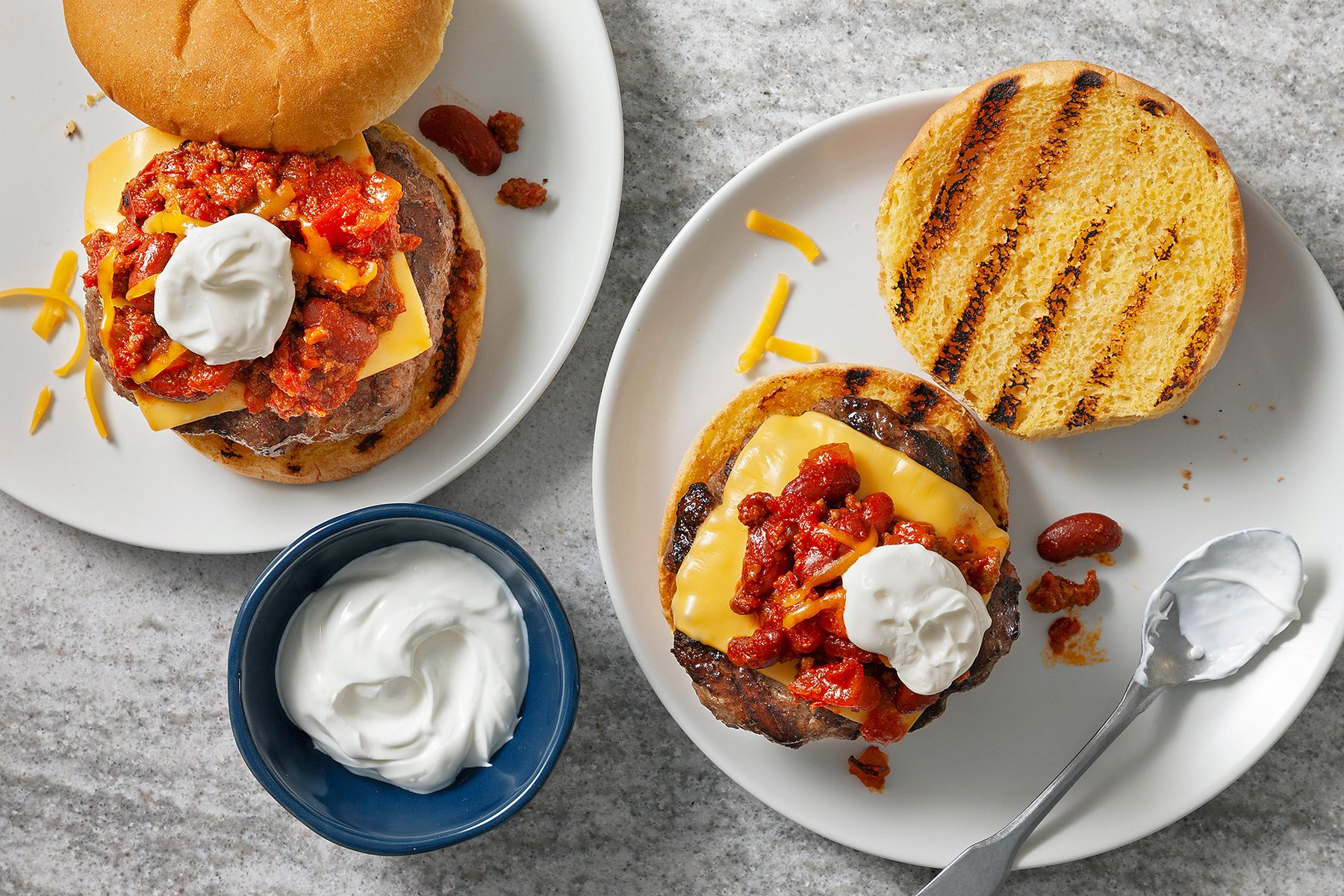 Two cheeseburgers with chili, cheese, and sour cream on buns are on separate plates. A small bowl of sour cream with a spoon is beside them on a gray surface.
