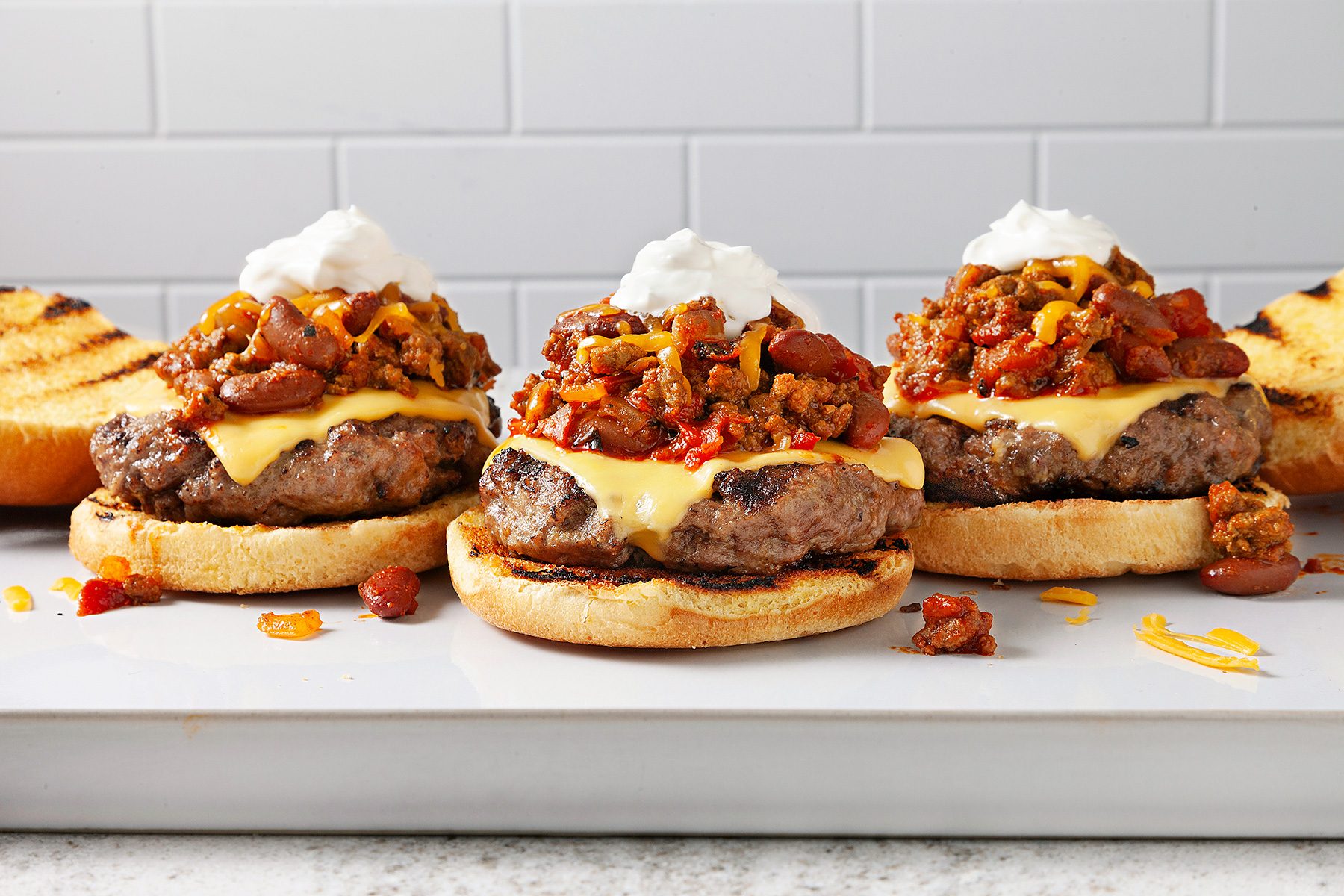 Three burgers on a plate with toasted buns are topped with melted cheese, chili with beans, shredded cheddar, and a dollop of sour cream. The background features a white tiled wall.