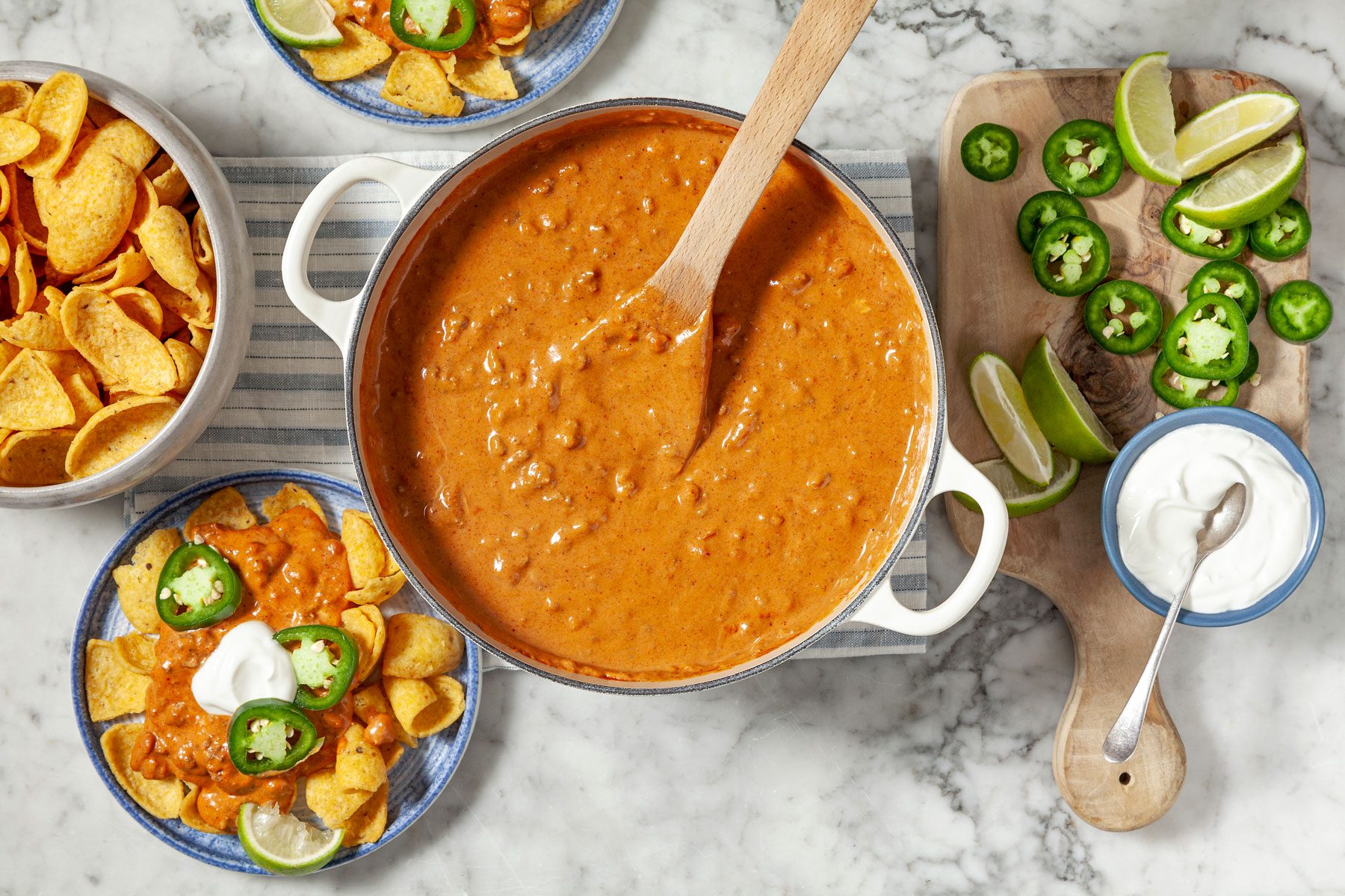 Chili served with corn chips. Sour cream, sliced jalapeno and lime wedges can be seen next to ithe bowl.
