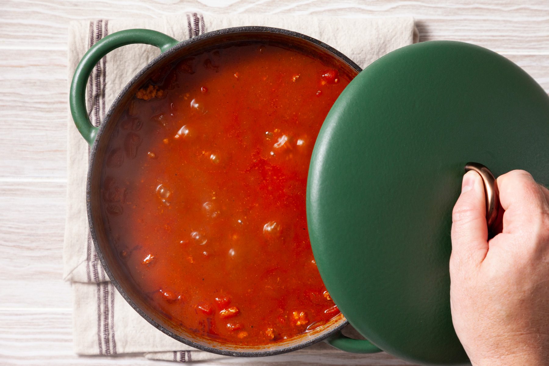 An overhead shot of a green cast-iron pot filled with simmering chili. A hand is lifting the lid, revealing a thick, red sauce with visible chunks of meat and vegetables. Steam rises from the pot, indicating that the chili is hot and ready to serve. The pot is placed on a wooden surface.