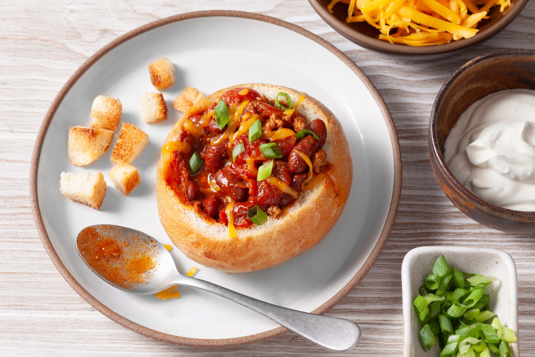 A 3/4 shot of a delicious and hearty chili in a bread bowl. A hollowed-out bread bun is filled with thick, red chili, topped with shredded cheese, chopped green onions, and a dollop of sour cream. A small bowl of croutons is served alongside the chili.