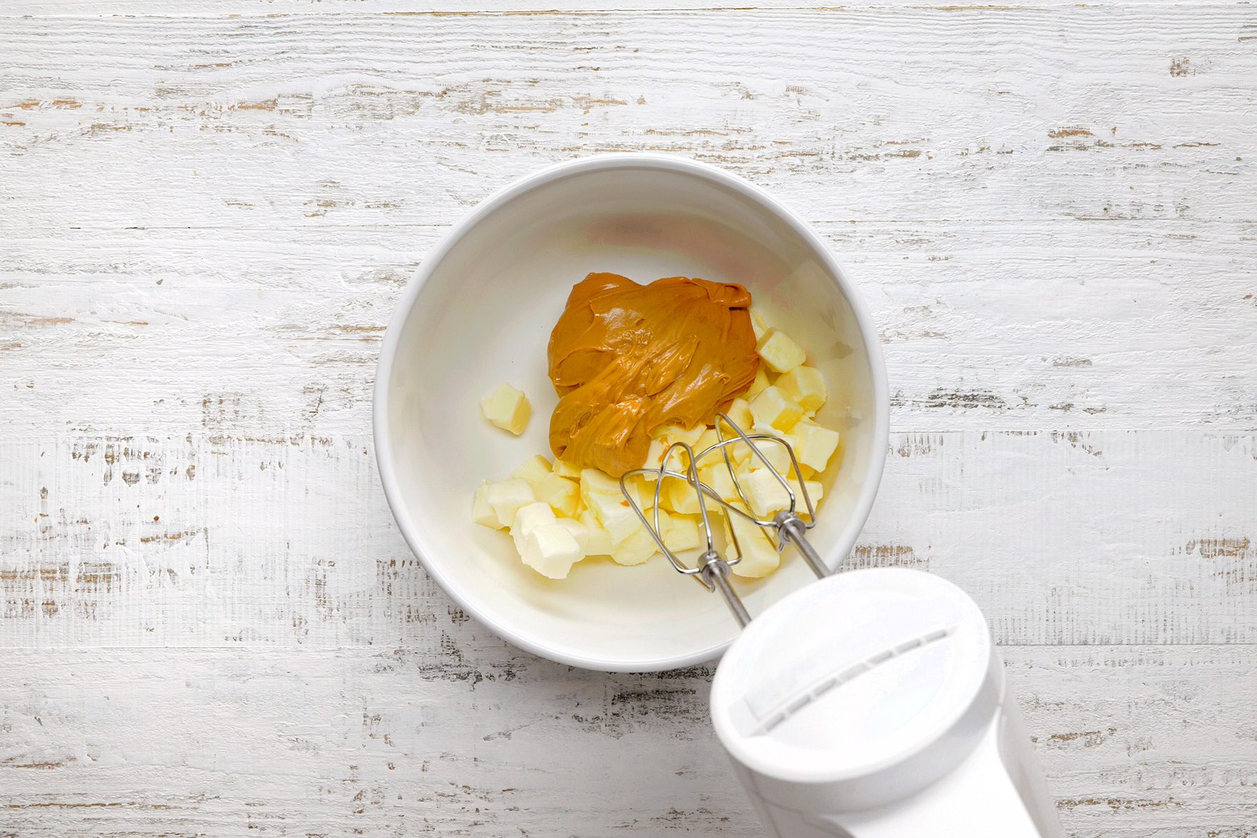A white mixing bowl contains chunks of butter and a dollop of peanut butter. A hand mixer with beaters is ready to blend the ingredients. The setup is on a rustic white wooden surface.