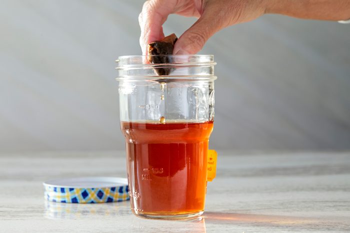 Horizontal shot of a hand dropping a tea bag into a glass jar with brewed tea. The jar, featuring a lid with a blue, yellow, and white design, contains deep amber tea with a small amount left. The tea bag is partially submerged, and the background is light gray.