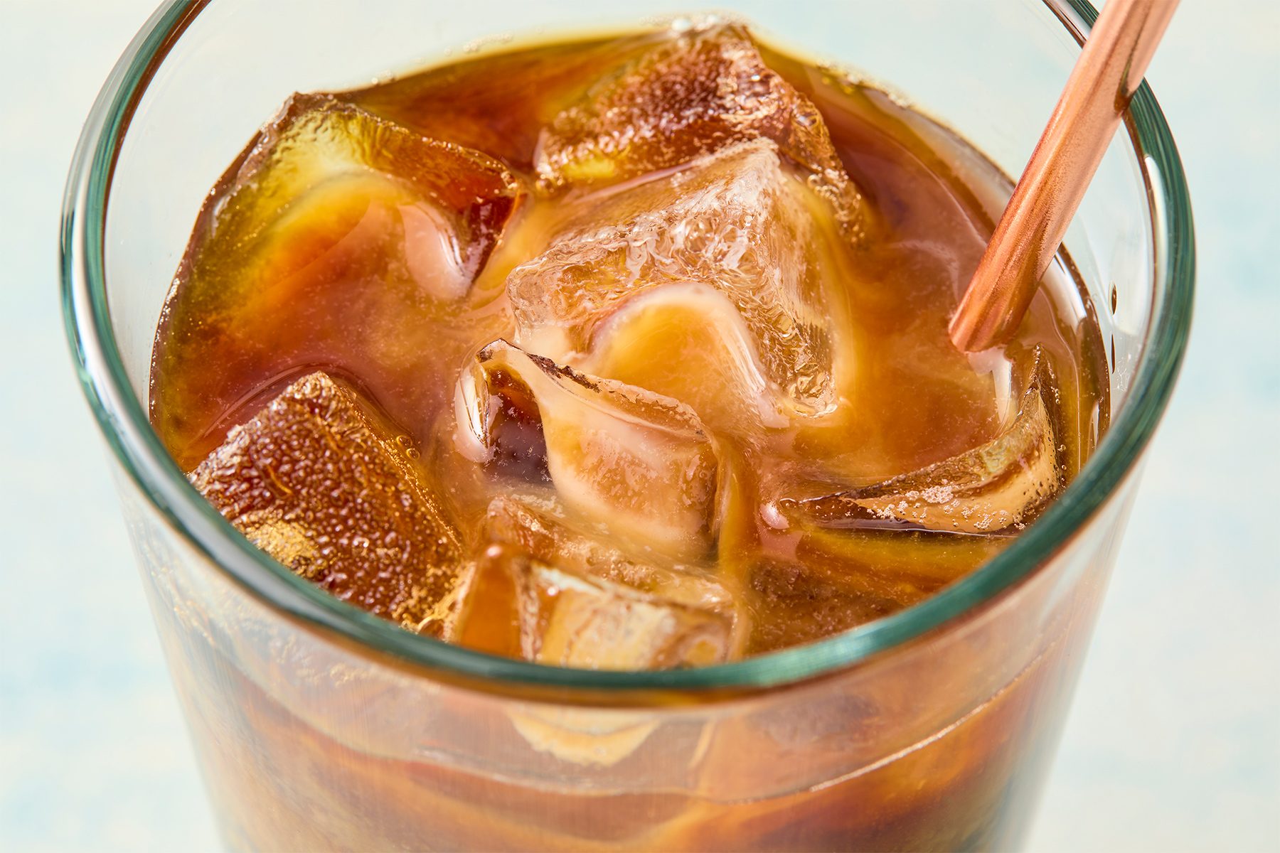 A glass of iced coffee with a brown straw. The coffee is dark and served over large ice cubes.