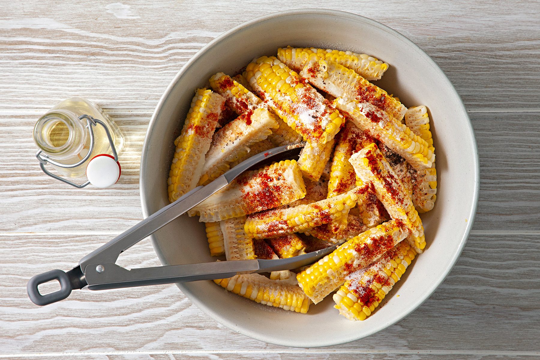 Overhead shot of place in a large bowl and toss in olive oil; In a small bowl mix together salt; garlic powder; onion powder; pepper; paprika and cayenne; sprinkle over corn and toss until evenly coated; tongs; wooden surface;