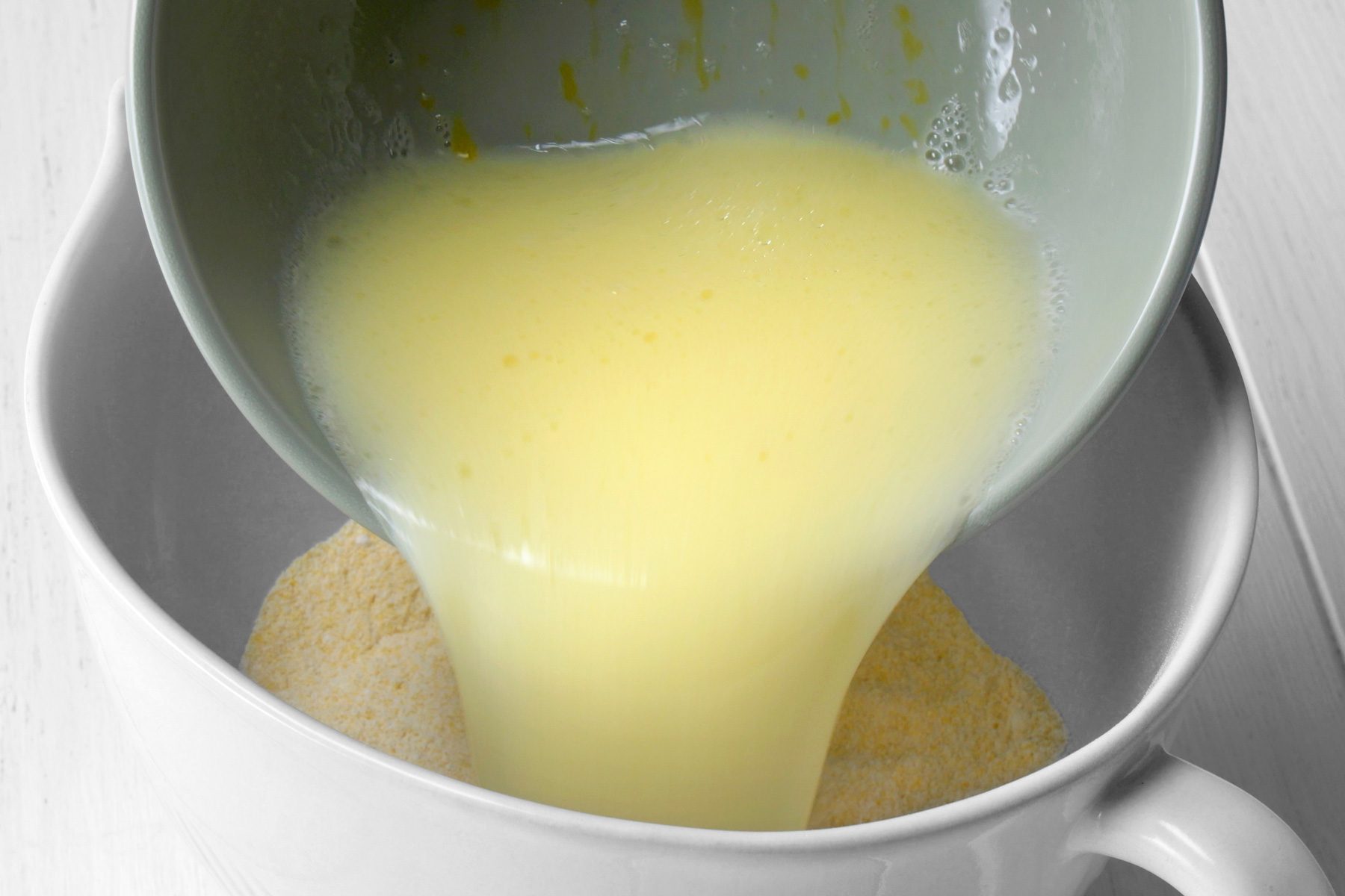 3/4th shot of a hand pouring a yellow liquid from a gray bowl into a larger white bowl, The liquid is pouring over a dry mixture in the white bowl, The dry mixture appears to be a light brown color and has a powdery texture;
