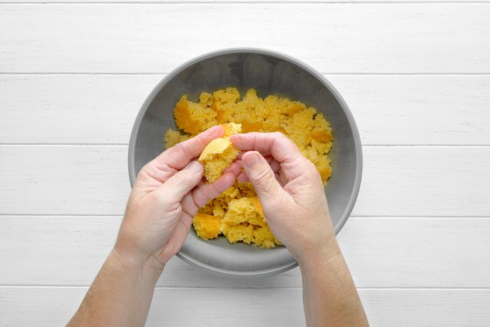 overhead shot of a hand tearing apart a piece of cornbread in a gray bowl, The cornbread appears to be golden brown and crumbly, The hand is breaking the cornbread into smaller pieces, The bowl is placed on a white wooden surface;