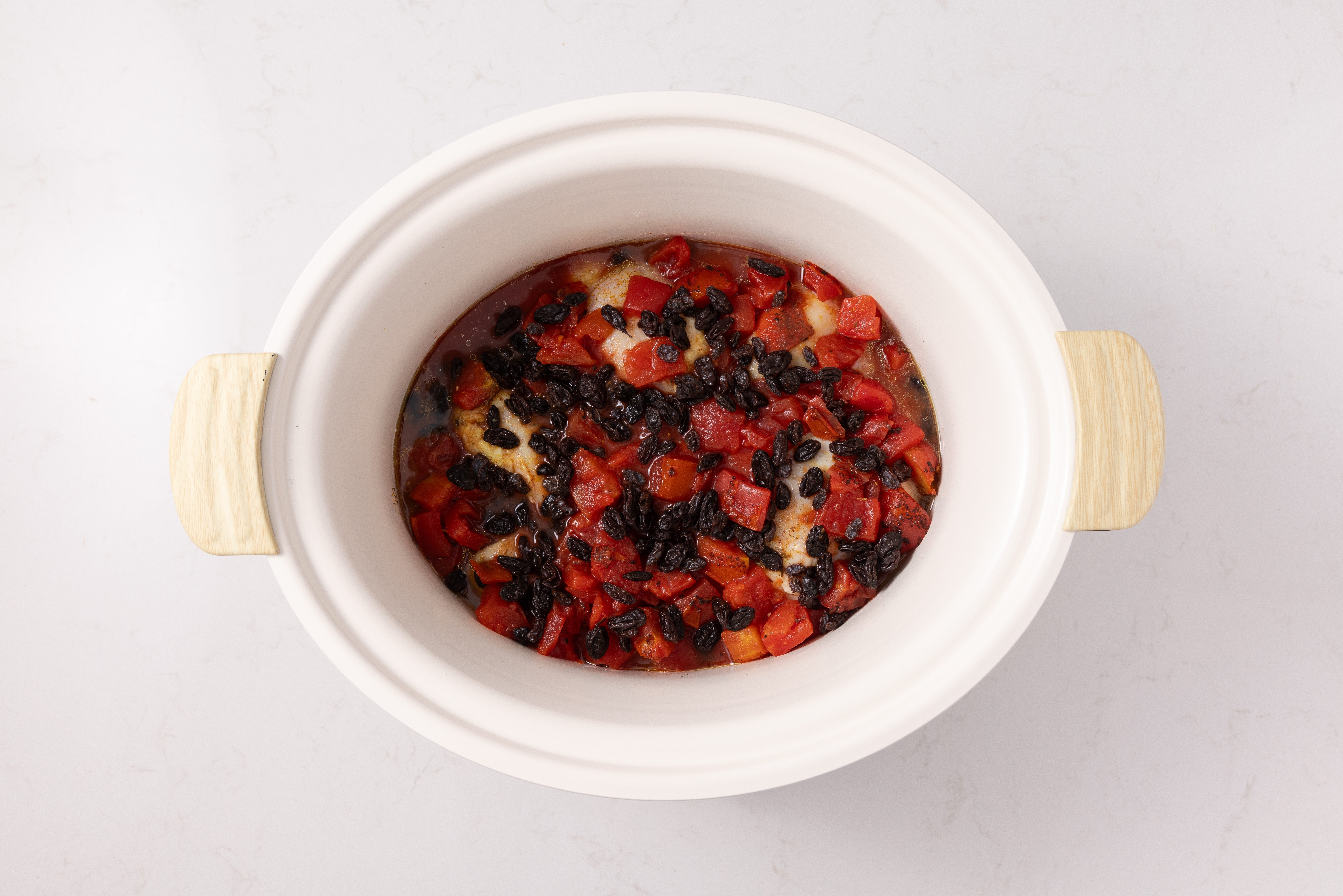 Chicken thighs cooking in slow cooker topped with diced tomatoes and raisins.