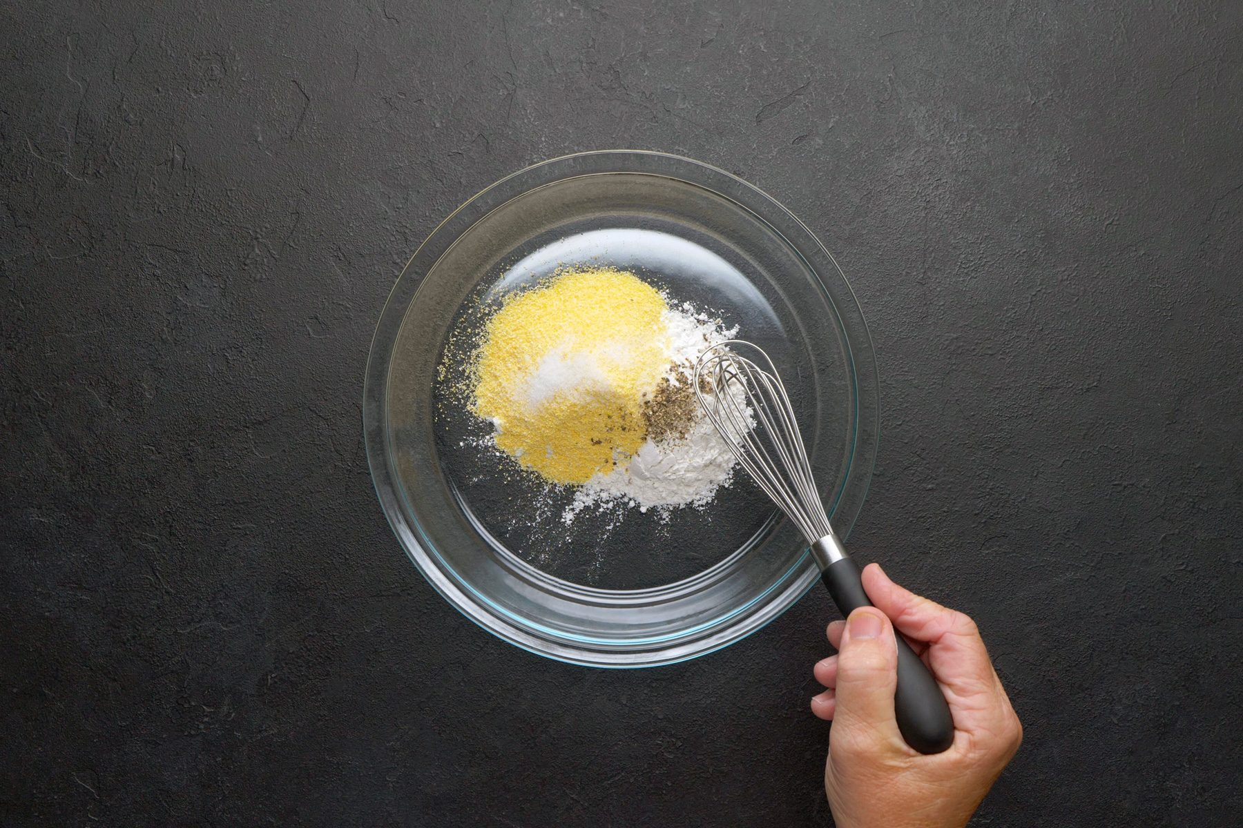 overhead shot of a glass bowl filled with a mixture of dry ingredients, The ingredients appear to be a light brown color and a white powder, A whisk is being used to stir the mixture together, The bowl is sitting on a black surface