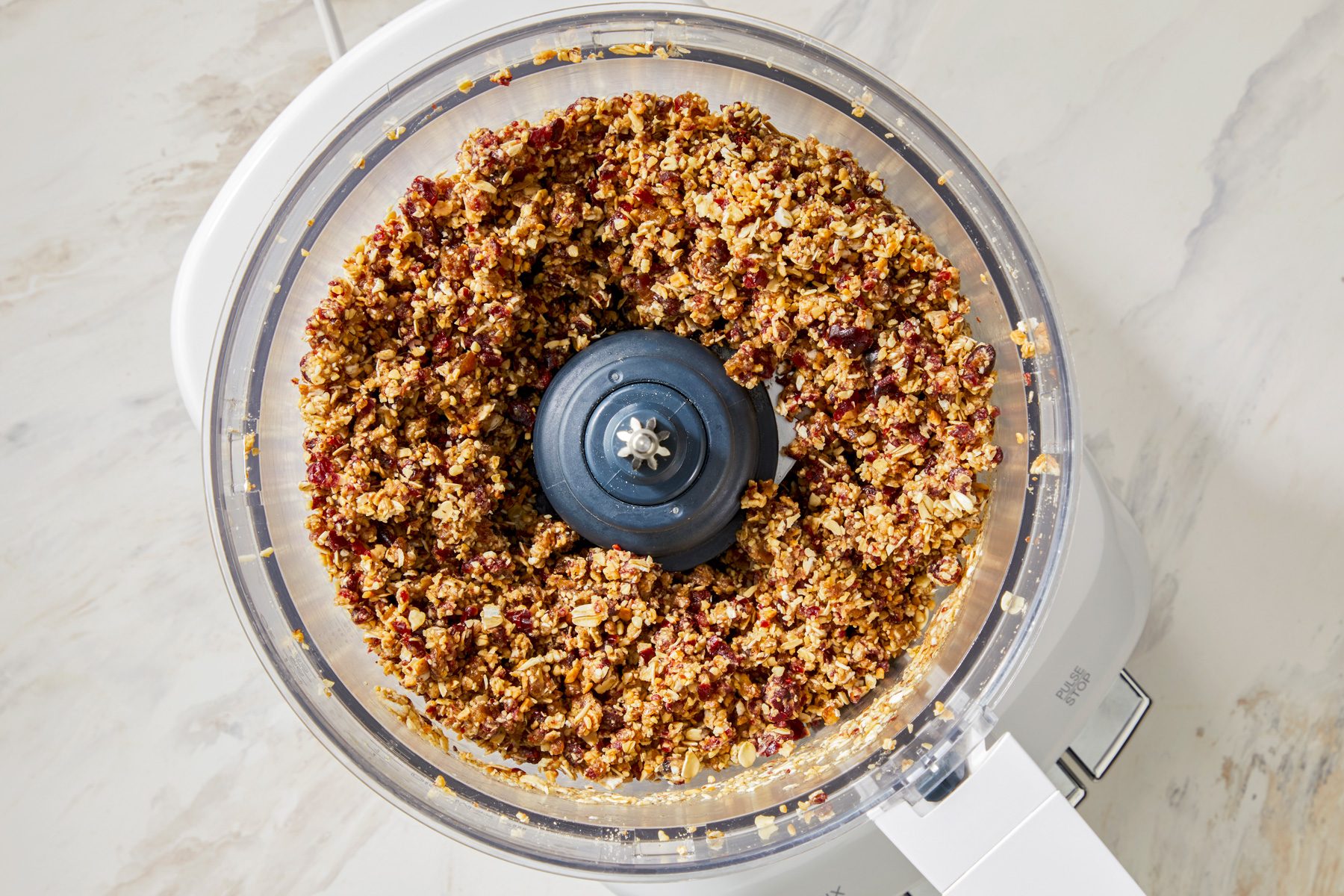 A food processor with the lid on, showing a mixture of oats, nuts, and dried fruits being pulsed.