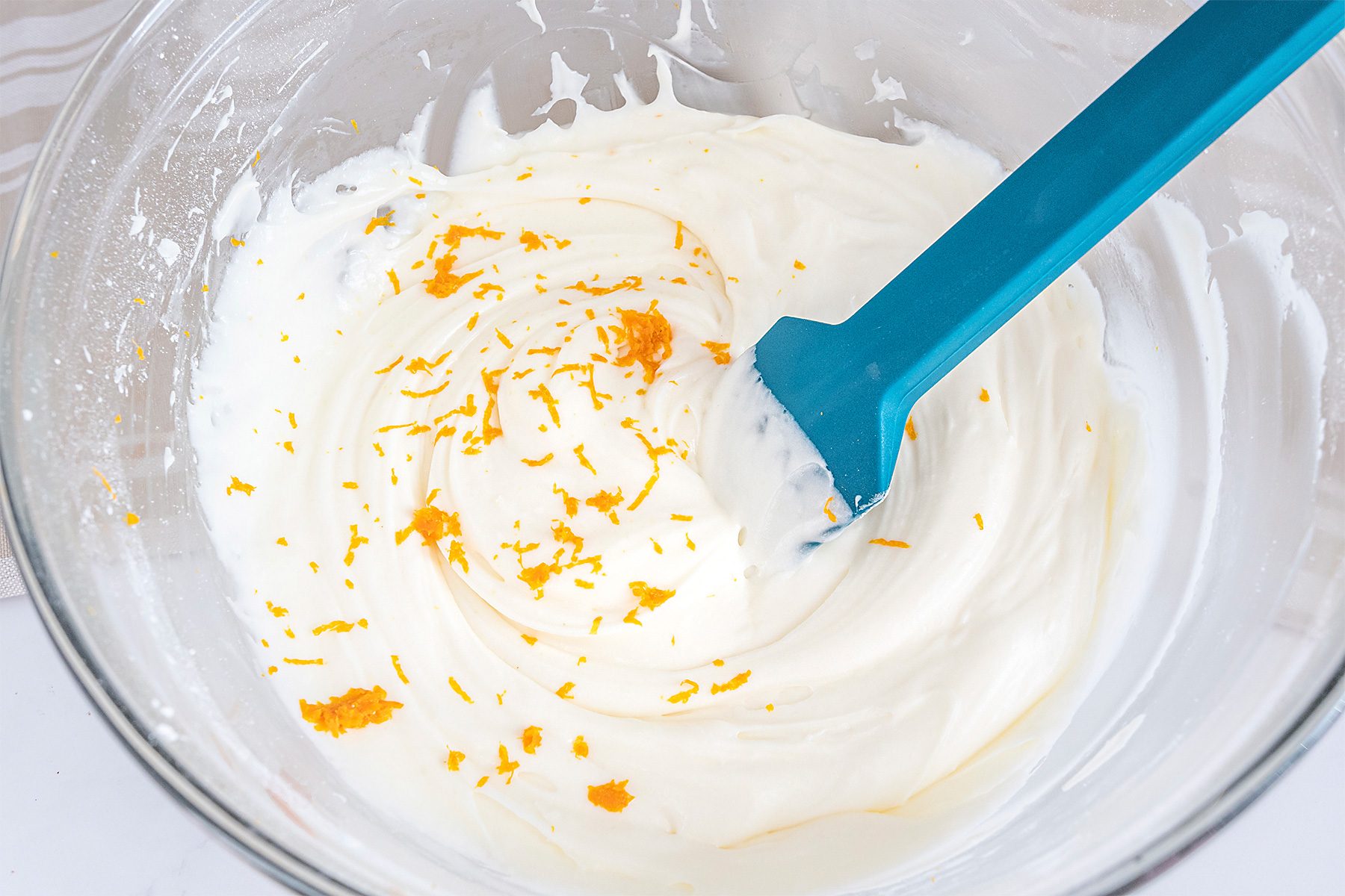 A glass bowl filled with a creamy white mixture is being stirred with a blue spatula. Orange zest is scattered on top of the mixture. The background is a light gray surface.