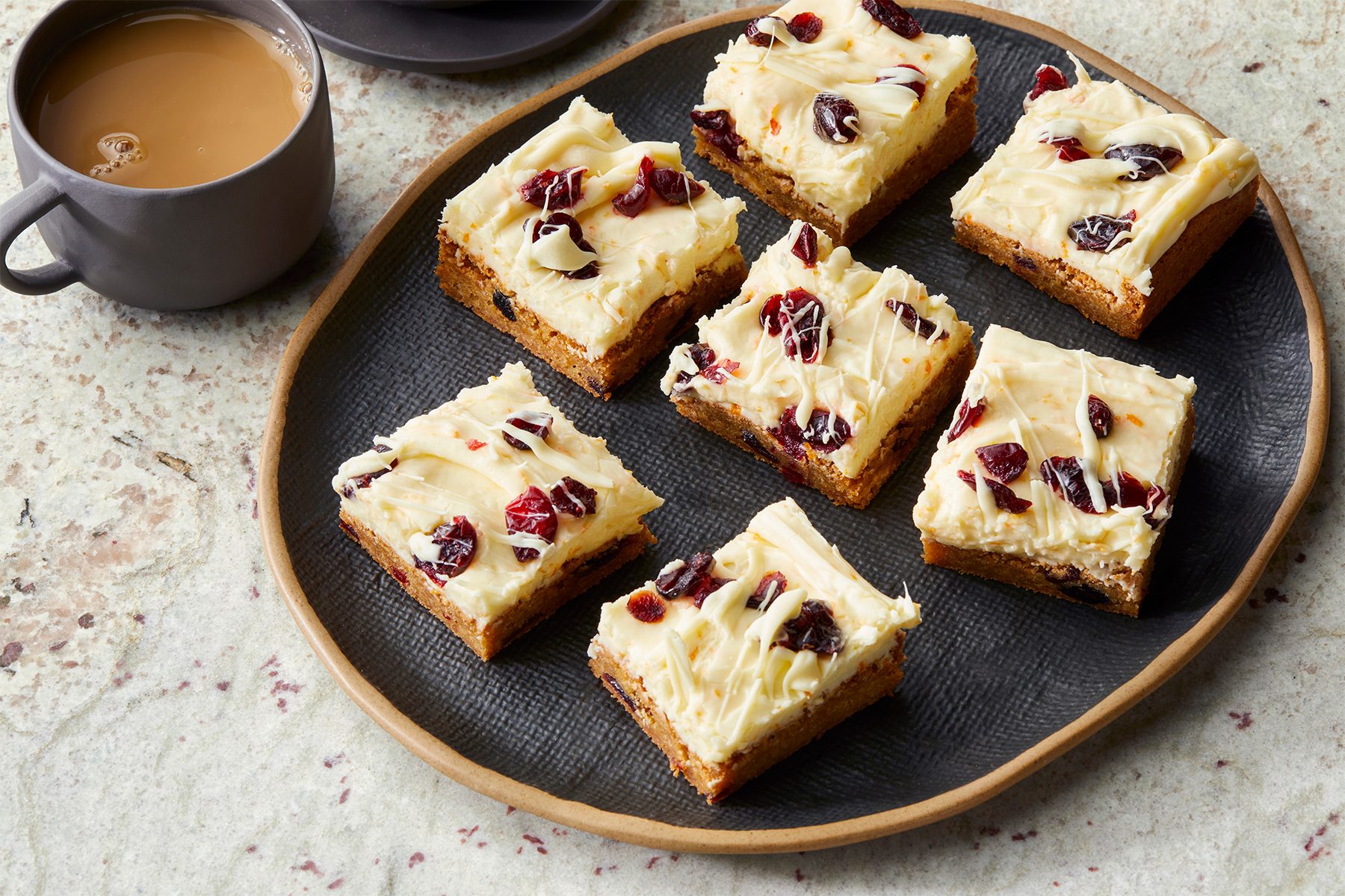 A dark plate holds eight square pieces of frosted bars topped with dried cranberries. Beside the plate is a cup filled with coffee or tea. The background is a textured, light-colored surface.