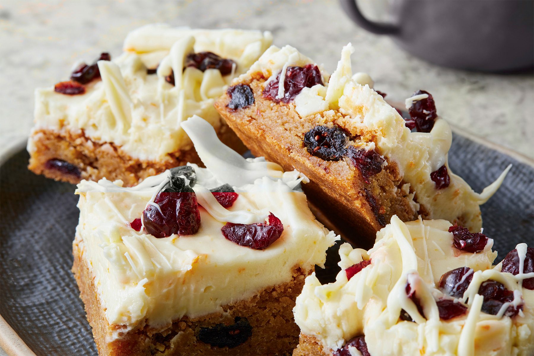 Four frosted blondies with dried cranberries and white chocolate shavings on top, served on a gray plate.