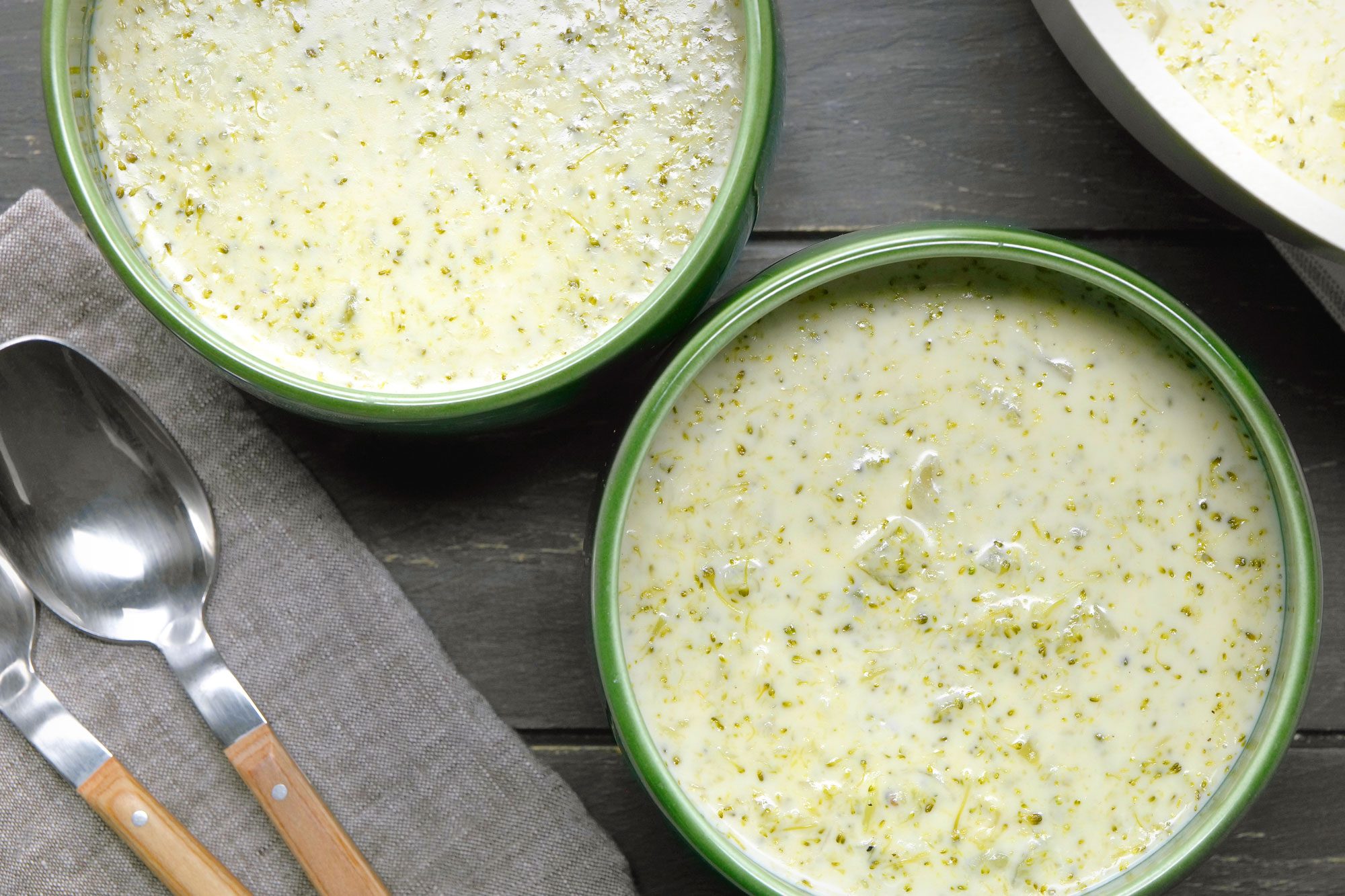 overhead shot of Two green bowls filled with Creamy Broccoli Soup are showcased, The soup is a creamy, pale yellow color with visible flecks of broccoli, The texture appears thick and smooth, Spoons are placed near the bowls;