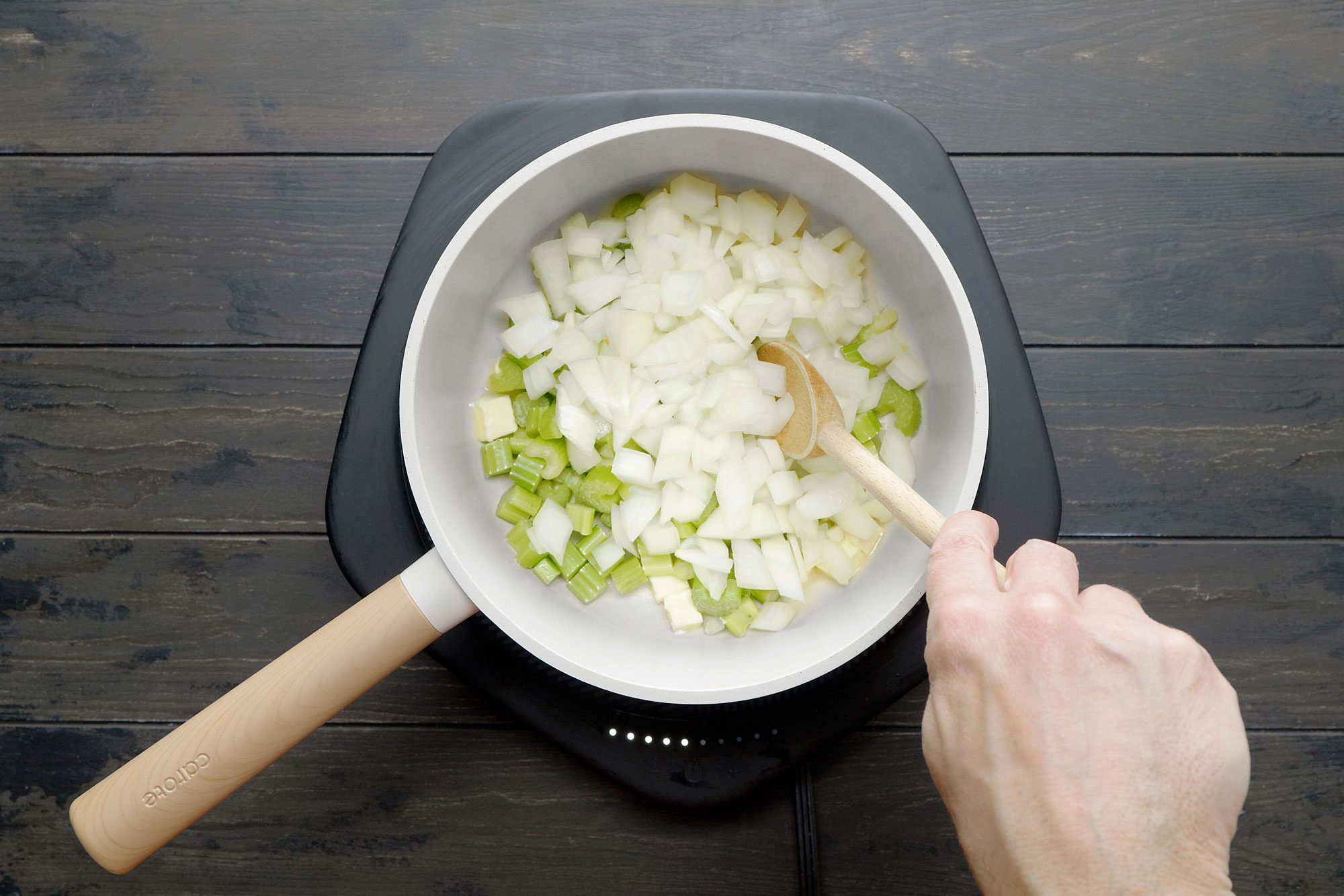 overhead shot of a white saucepan is placed on an electric induction cooktop, The saucepan contains chopped onions and celery, which are being stirred with a wooden spoon, The onions are white and the celery is green; the vegetables are being sautéed;