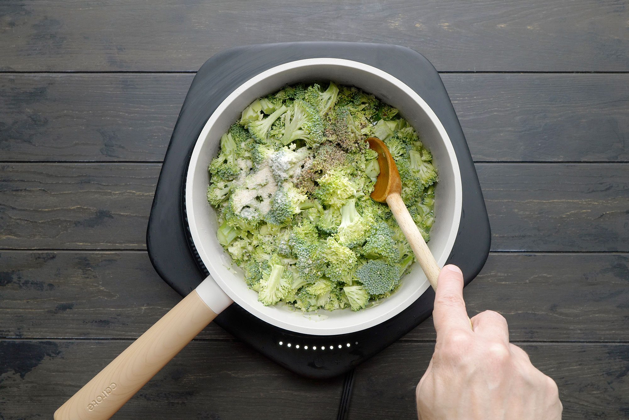 overhead shot of a white saucepan is filled with chopped broccoli florets, A wooden spoon is stirring the broccoli, and a sprinkle of seasonings, likely salt and pepper, is visible on top, the broccoli is being cooked and seasoned;