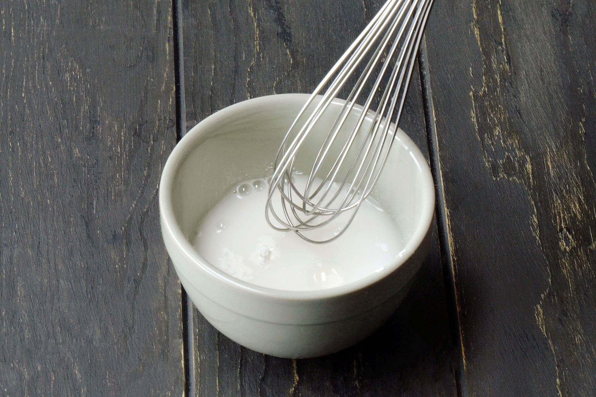 3/4th shot of a white bowl contains a liquid mixture that is being whisked with a metal whisk, The liquid appears to be a light yellow color and has bubbles forming on the surface, The bowl is placed on a black wooden surface;