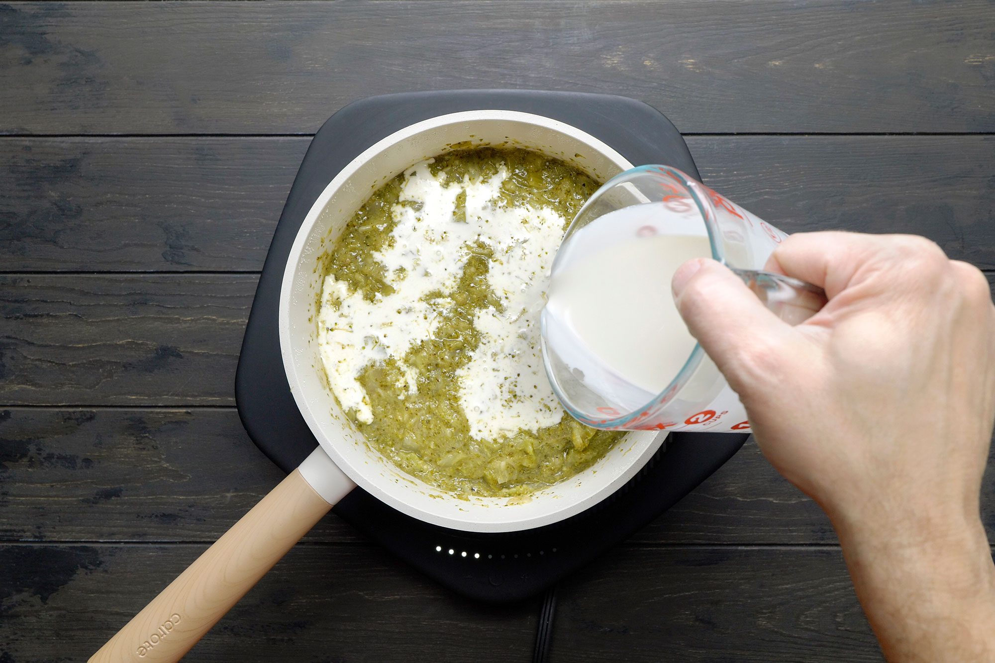 overhead shot of a white saucepan on an electric induction cooktop contains a creamy, green mixture, A person's hand is pouring a liquid, likely cream or milk, into the saucepan, The liquid is creating ripples and swirls in the mixture;