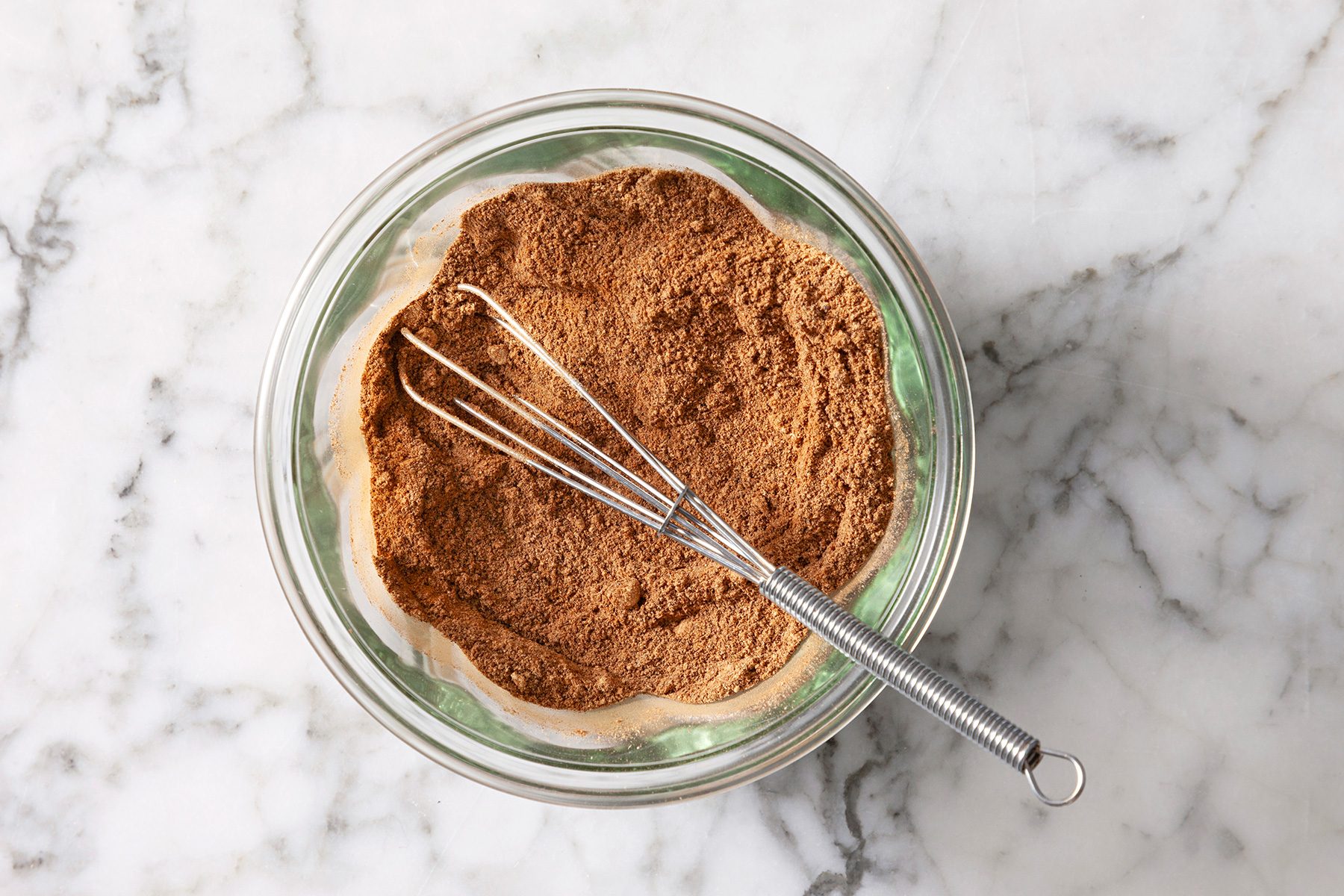 A clear glass bowl filled with brown spice mix, placed on a marble surface. A metal whisk rests inside the bowl.
