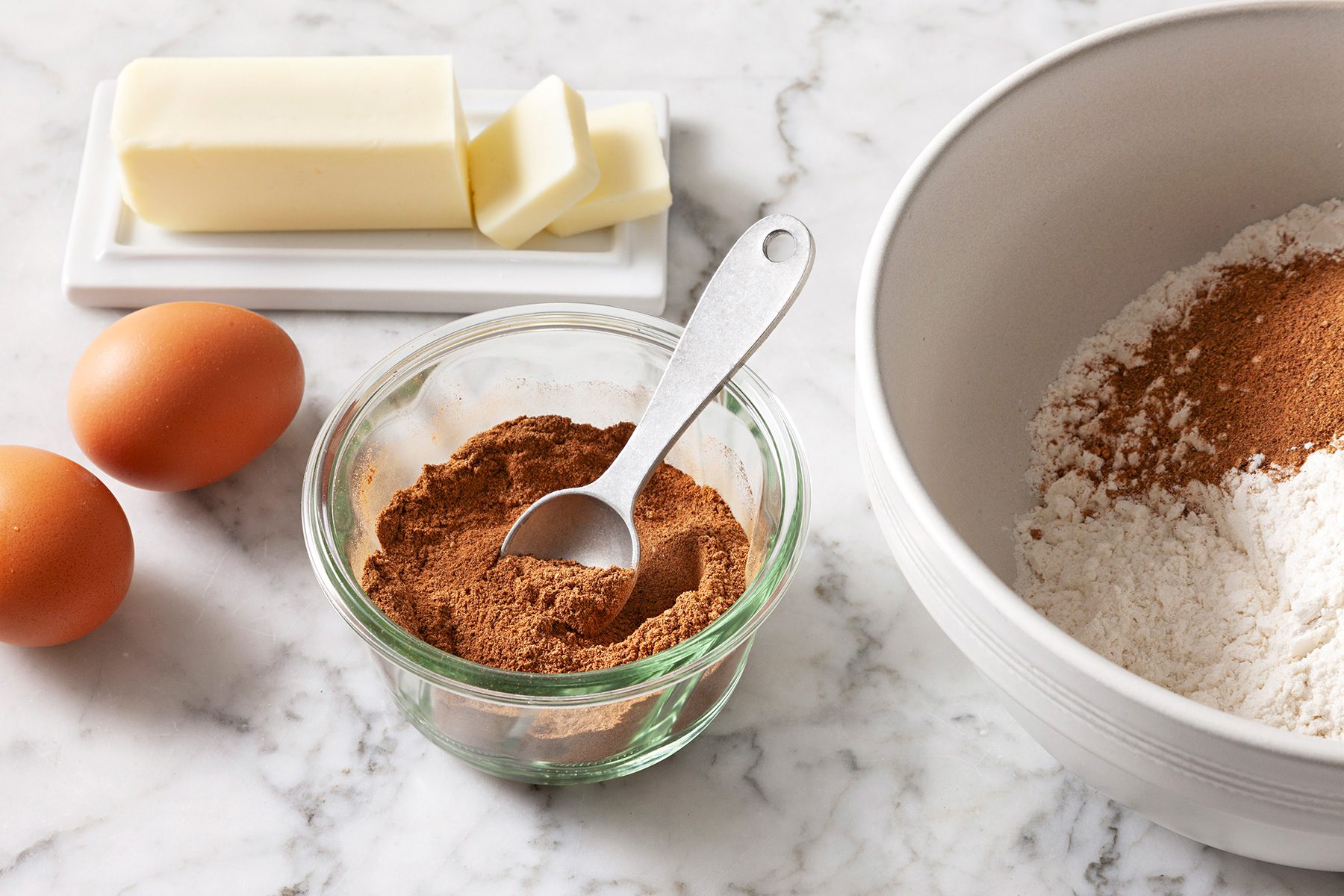 Baking ingredients on a marble countertop: two eggs, a stick of butter on a dish, a small jar of brown powder with a spoon, and a mixing bowl containing flour and more brown powder.