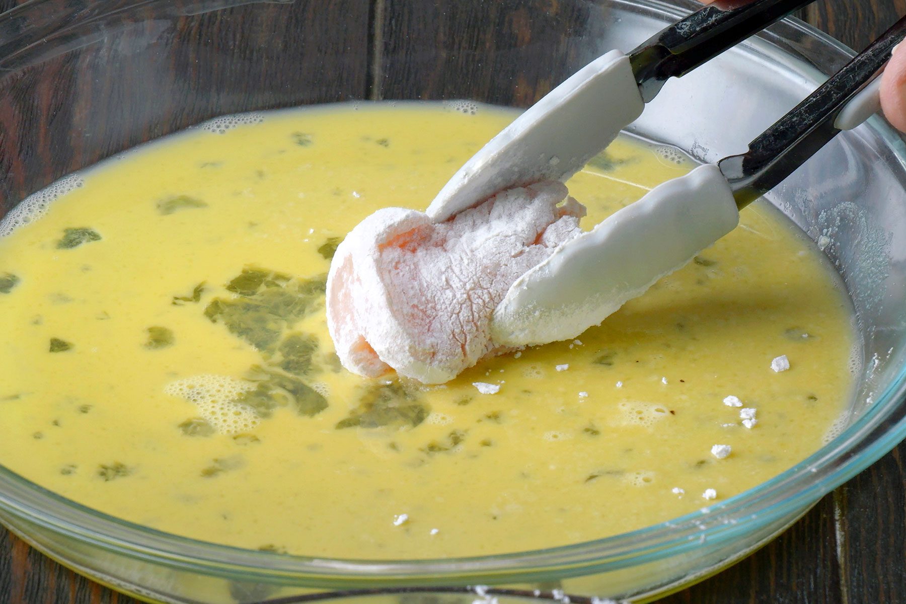 Tongs holding a flour-coated piece of chicken being dipped into a yellow, herb-spiced batter in a clear glass bowl on a wooden surface.