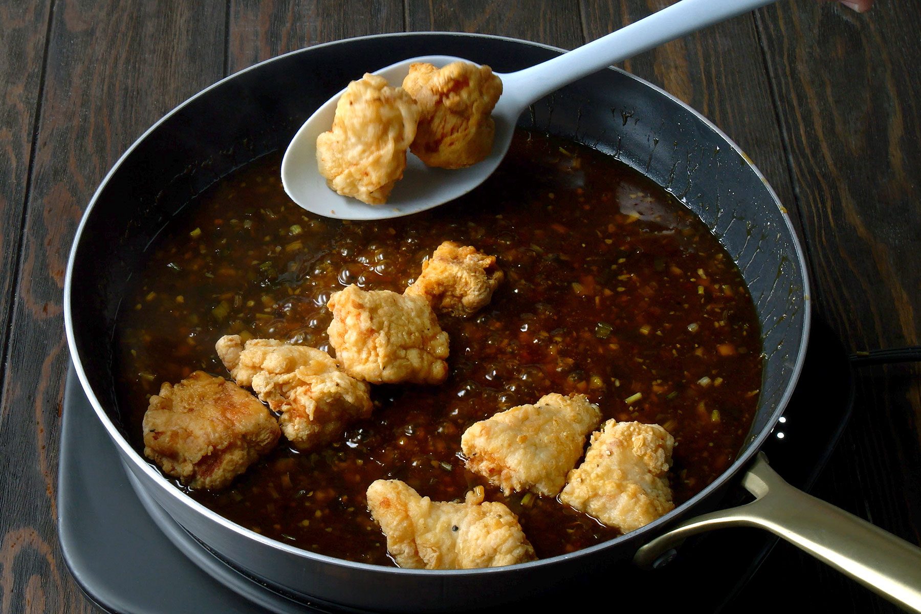Fried chicken pieces being scooped from a pan filled with a thick, dark sauce. The sauce seems to be a savory glaze with visible spices and small ingredients. The pan is on a dark wooden surface.