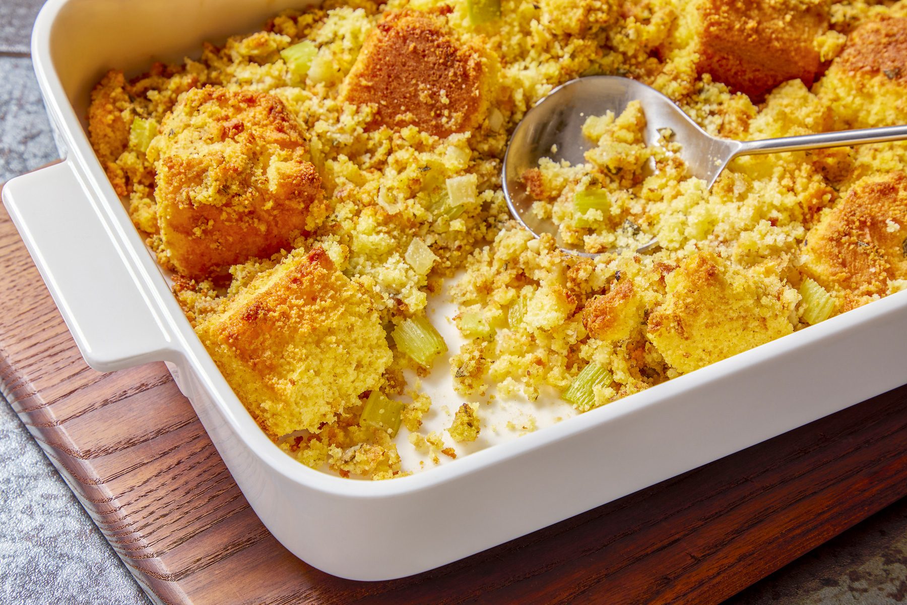 3/4th shot of a white baking dish filled with golden brown cornbread stuffing is sitting on a wooden cutting board, The stuffing is dotted with small pieces of celery and sage, A spoon is laying in the upper left hand corner of the photo, Behind the cutting board, a white plate with a green napkin is in the lower right corner, The background is a dark gray speckled countertop.