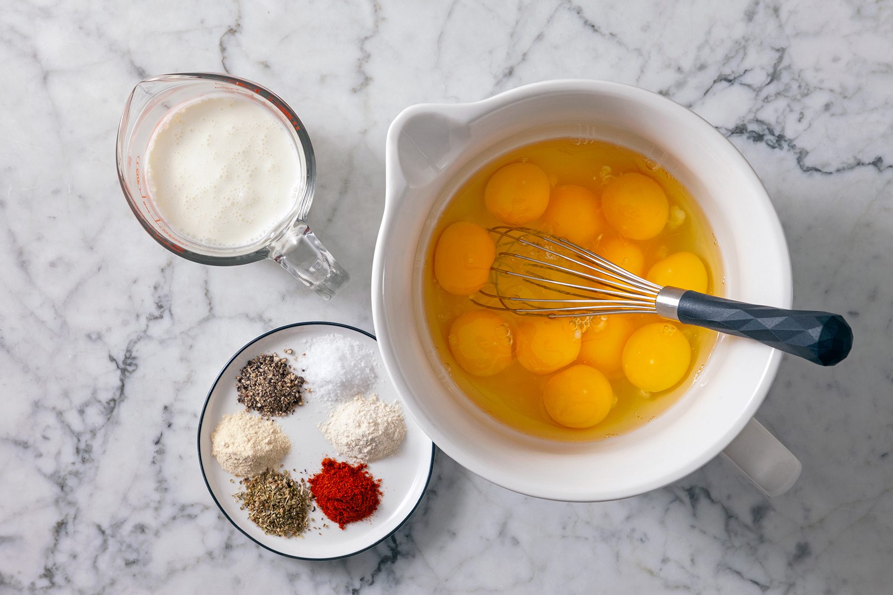 A white bowl contains several cracked eggs and a whisk. A small pitcher of milk sits nearby. A plate with various spices, including black pepper, salt, flour, oregano, and paprika, is also on the marble countertop.