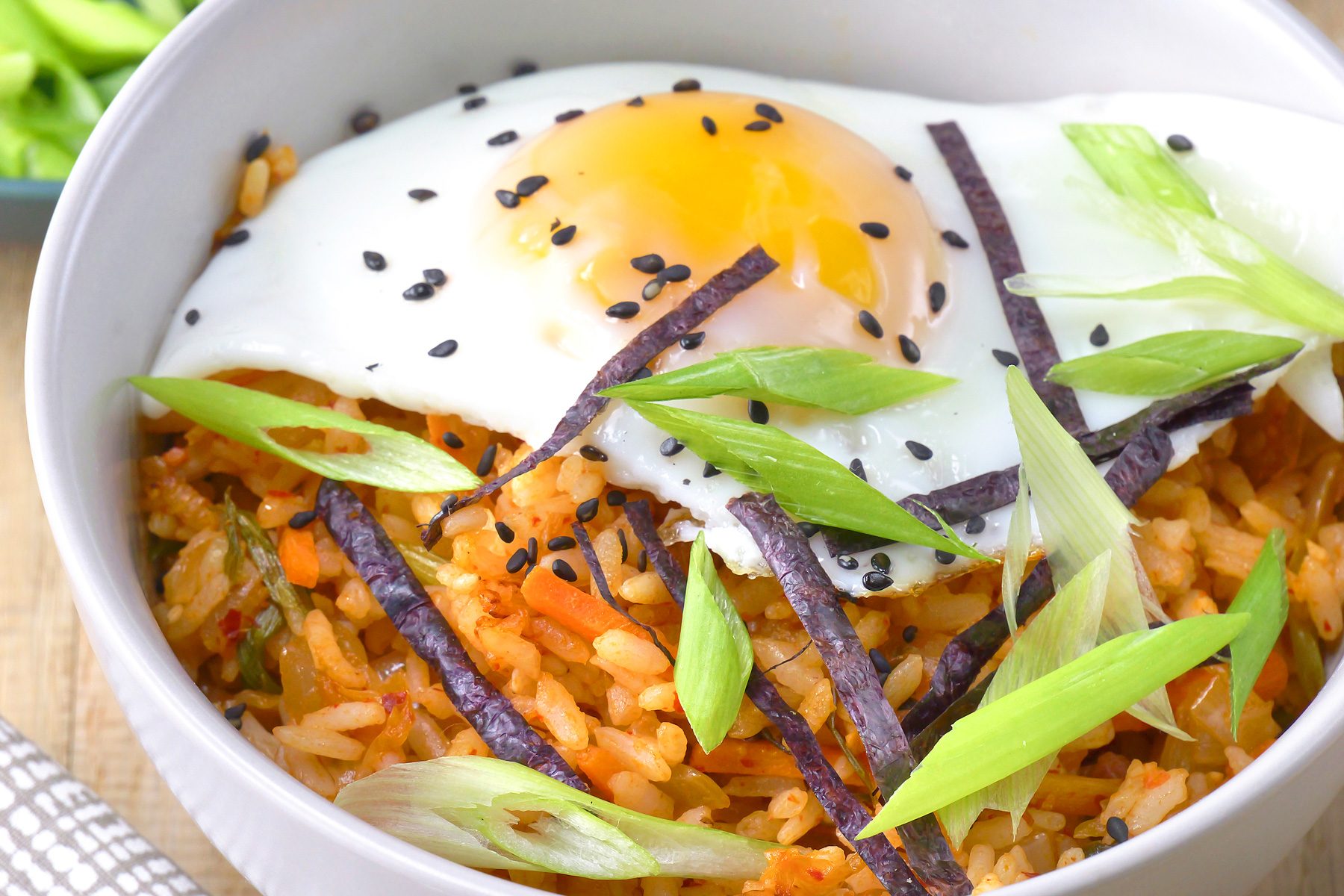 close shot of a bowl of Kimchi Fried Rice is showcased, featuring a vibrant mixture of rice, kimchi, vegetables, and a fried egg on top, The rice is tinged with a reddish hue from the kimchi;