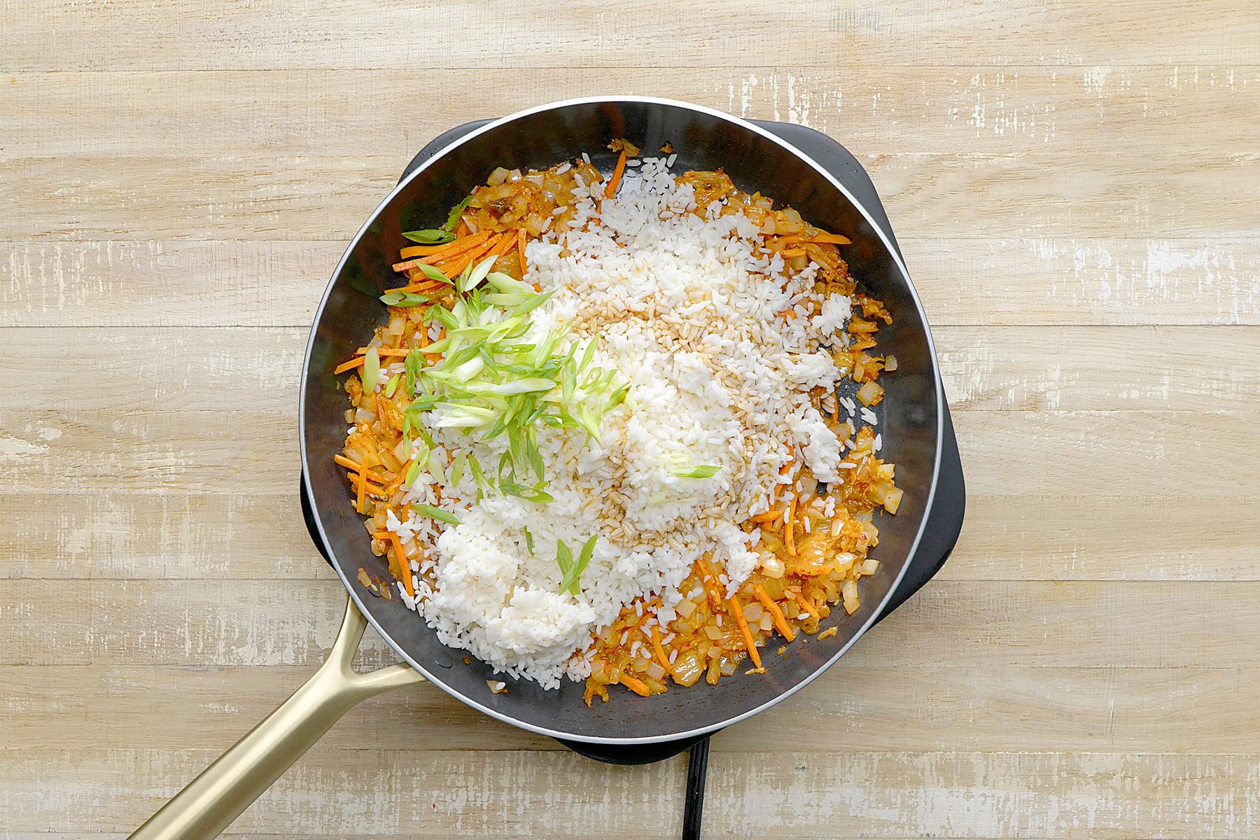 overhead shot of a black skillet contains a mixture of sautéed vegetables, kimchi, and cooked rice, The vegetables are a vibrant orange and white color, and the rice is a white color, Chopped green onions are scattered on top of the mixture;