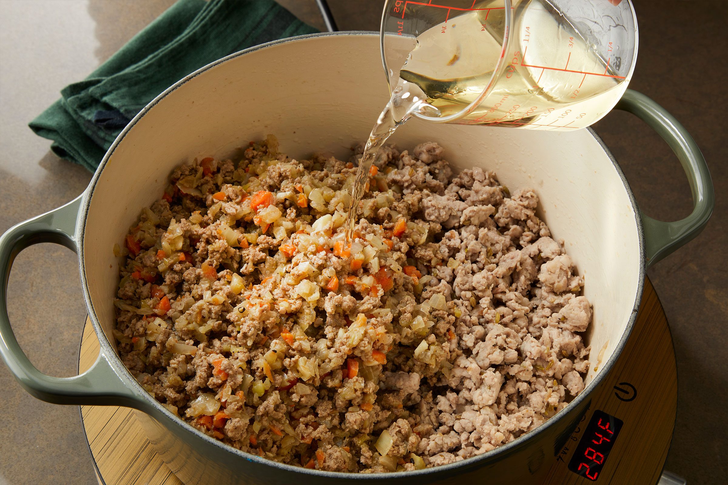 Adding broth to crumbles of sausage and beef with vegetables to prepare Pappardelle Bolognese in a pan