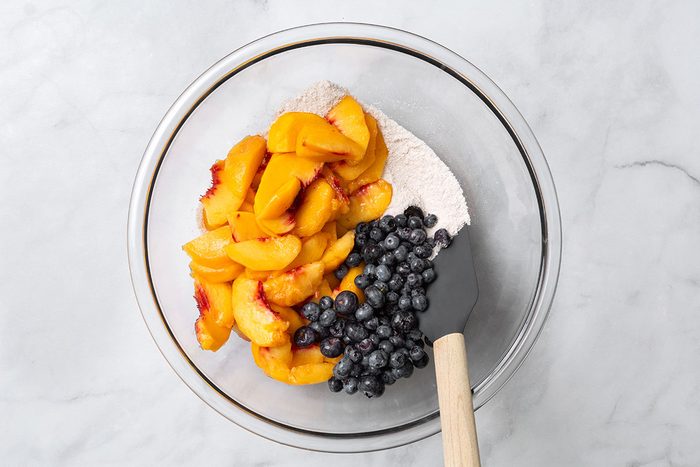 Sugar, flour, cinnamon and allspice in a mixing bowl with added sliced peaches and blueberries for step one of Peach and Blueberry Pie for Taste of Home