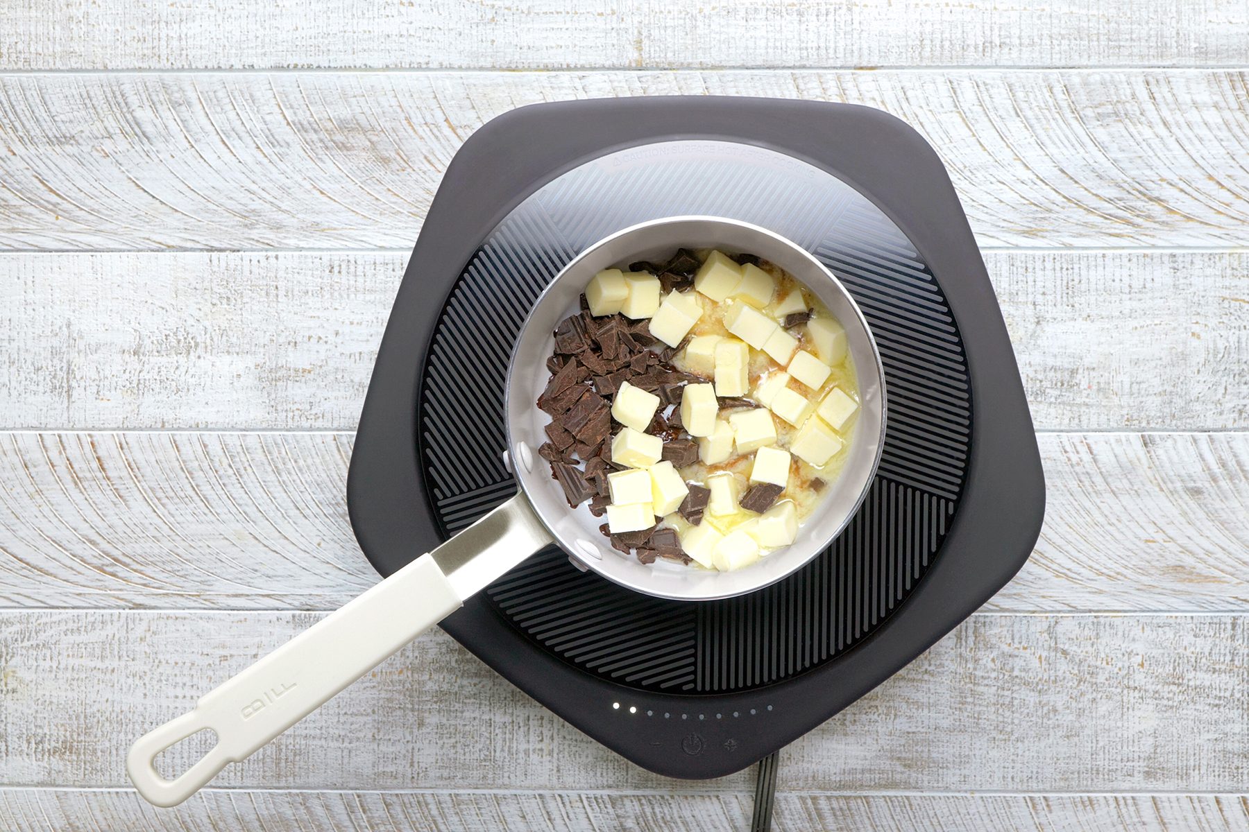 A stovetop with a saucepan containing chopped dark and white chocolate, ready for melting. The surface is wood-textured, and the setup rests on an electric cooktop.