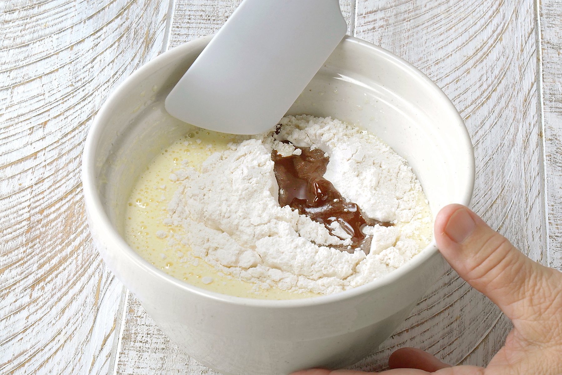 A person holding a white bowl filled with flour and a dark brown liquid, using a spatula to mix the ingredients. The bowl is placed on a light wooden surface.
