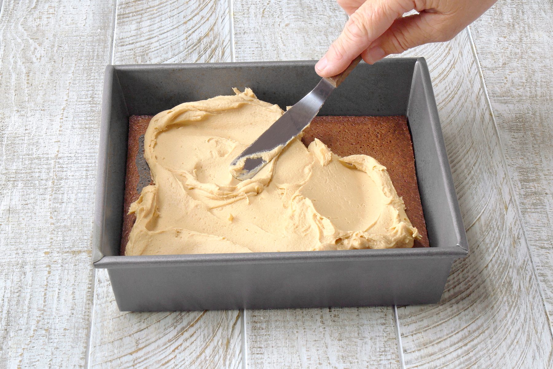 A hand uses a metal spatula to spread creamy frosting over a brown cake in a square metal baking pan on a textured white wooden surface.
