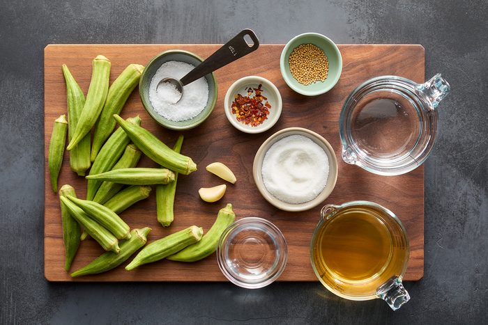 A wooden board displaying fresh okra pods, bowls of salt, pepper flakes, mustard seeds, and sugar, two garlic cloves, measuring spoons, a glass of water, and a glass of vinegar. Ingredients are neatly arranged for a recipe.