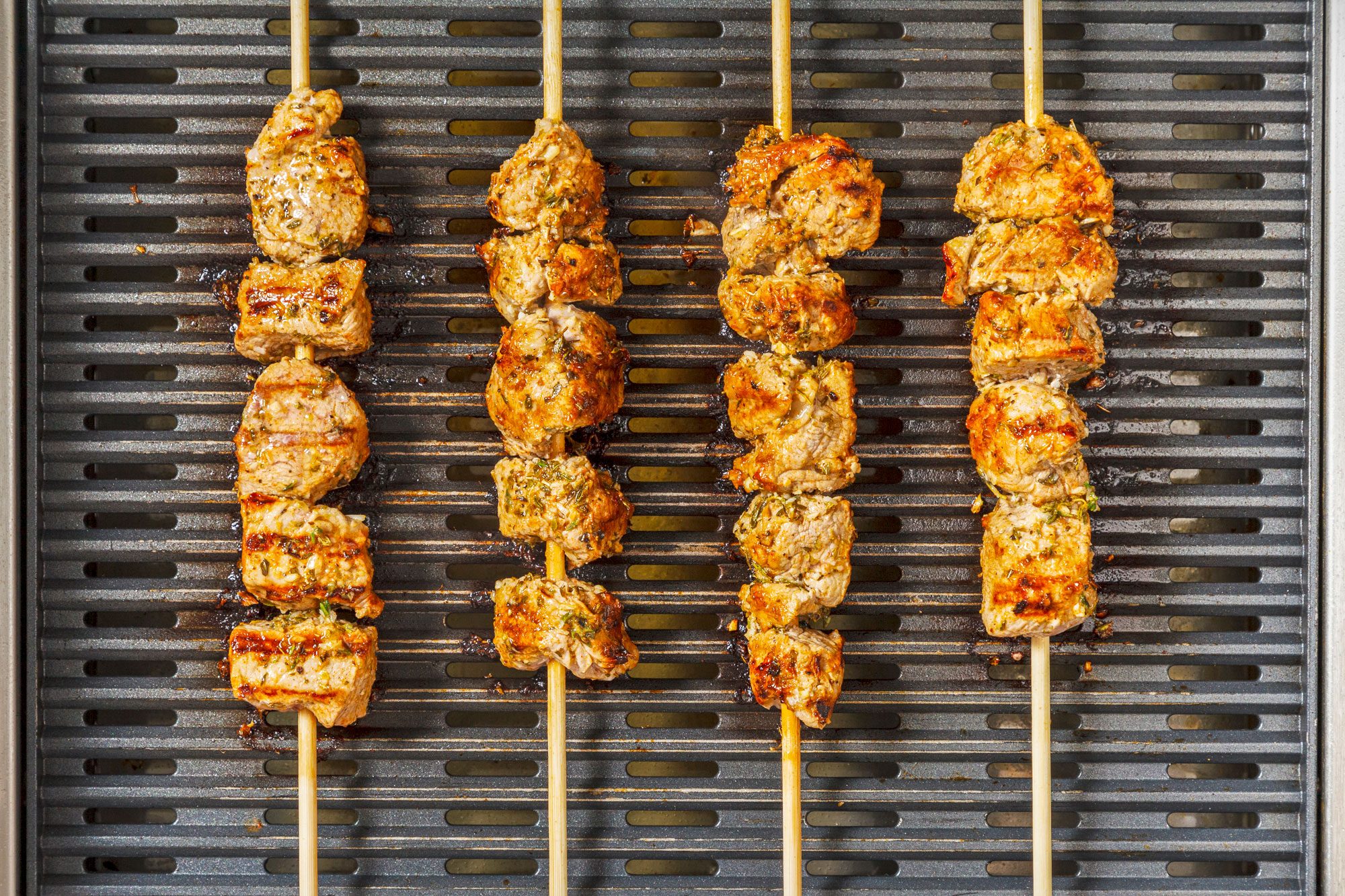 overhead shot of four skewers of grilled meat on a grill, The meat is cooked to a golden brown color and is slightly charred, The skewers are arranged in a parallel pattern on the grill;