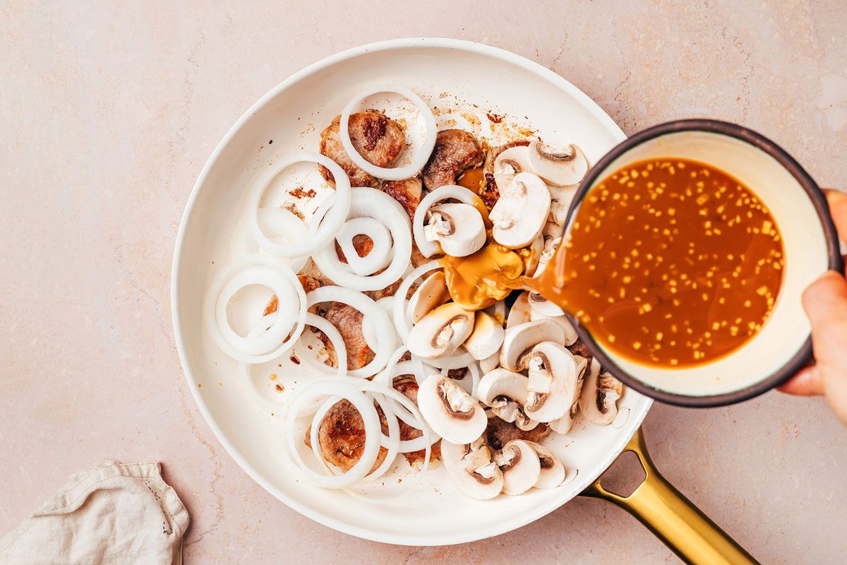 Brown Gravy mixture being poured in the skillet with cooked pork tenderloin, onion and mushrooms for the Pork Tenderloin Recipe by Taste of Home